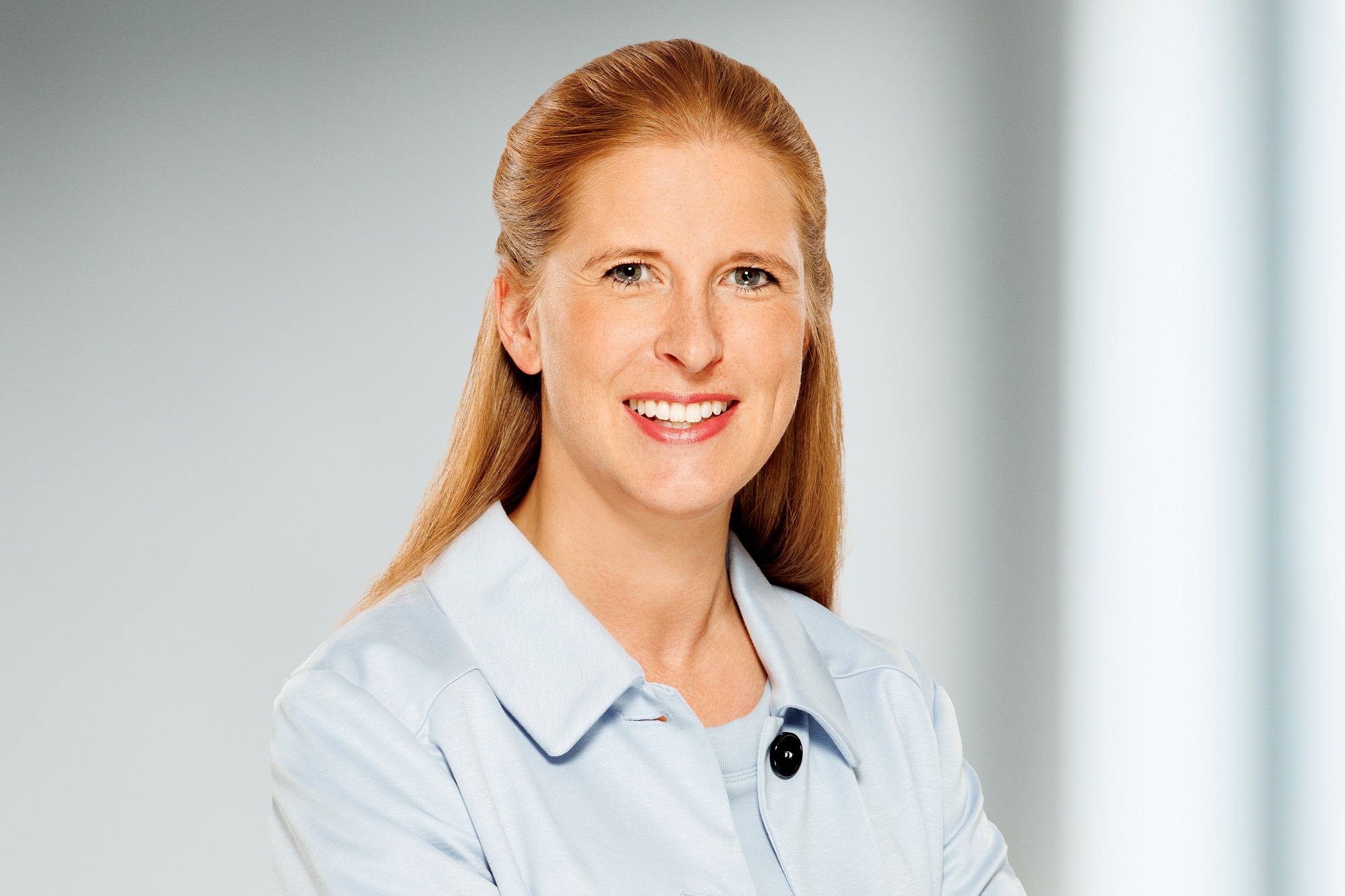 A woman with long, light brown hair smiles warmly, wearing a light blue jacket over a sweater. She stands confidently with her arms crossed, against a soft, neutral background that emphasizes her cheerful expression.