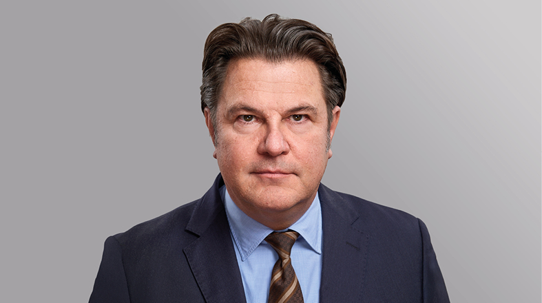 Portrait eines Mannes mit kurzen, leicht gewellten Haaren, der einen dunklen Anzug und ein helles Hemd trägt. Er hat einen ernsthaften Ausdruck und blickt direkt in die Kamera. Der Hintergrund ist grau und unscharf.