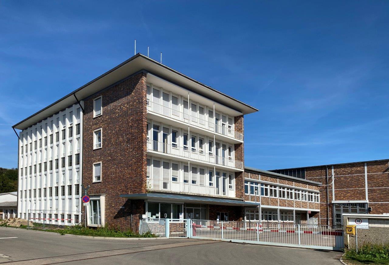 Das Bild zeigt ein modernes, dreigeschossiges Gebäude mit einer Kombination aus roten Ziegeln und weißen Fenstern. Im Vordergrund sind ein Zaun und eine Zufahrtsstraße sichtbar. Der Himmel ist blau und klar, was auf einen sonnigen Tag hinweist.