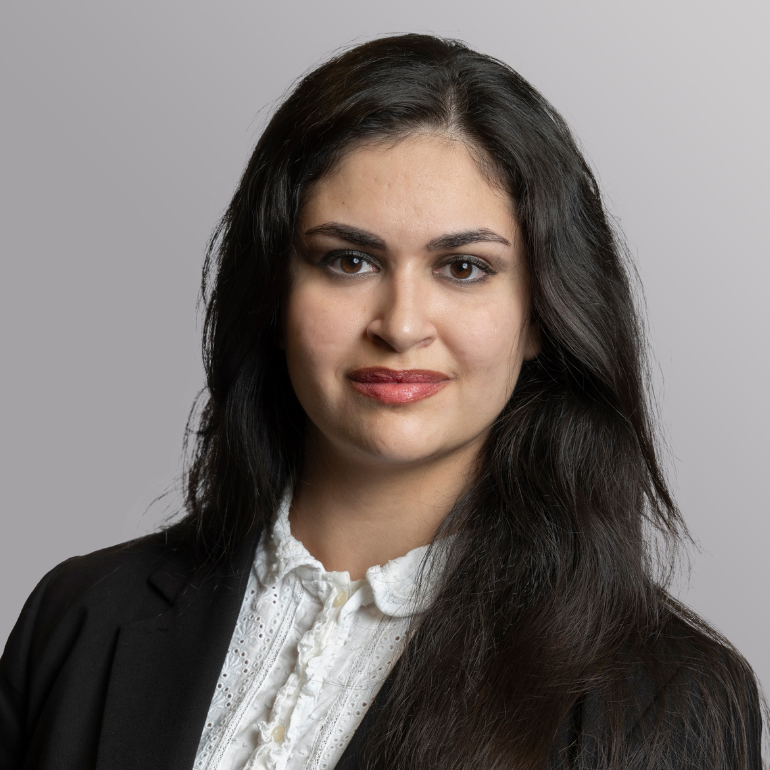 Ein Porträt einer Frau mit langen, dunklen Haaren. Sie trägt eine schwarze Jacke und ein weißes Hemd mit feinen Mustern. Ihr Gesichtsausdruck ist freundlich und selbstbewusst, während der Hintergrund in dezenten Grautönen gehalten ist.