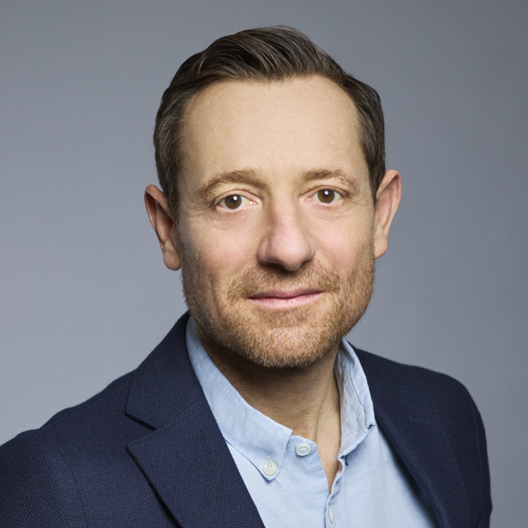 Ein lächelnder Mann mit kurzen, braunen Haaren und einem gepflegten Bart. Er trägt ein hellblaues Hemd und einen dunklen Blazer. Der Hintergrund ist einfarbig grau.