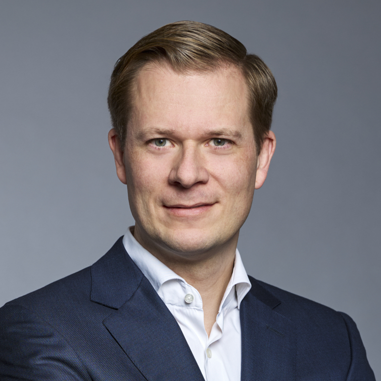 Porträt eines Mannes mit kurz geschnittenem, blonden Haar, der in einem dunkelblauen Anzug und einem weißen Hemd gekleidet ist. Er lächelt leicht, während der Hintergrund grau ist.