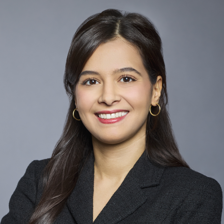 Porträt einer lächelnden Frau mit langen, dunklen Haaren, die einen schwarzen Blazer trägt. Der Hintergrund ist grau. Sie strahlt Freundlichkeit und Selbstbewusstsein aus.