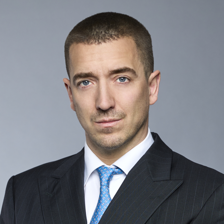 Ein ernst blickender Mann mit kurzgeschorenen Haaren trägt einen dunklen Anzug und eine hellblaue Krawatte. Der Hintergrund ist grau. Sein Gesichtsausdruck ist selbstbewusst und neutral.