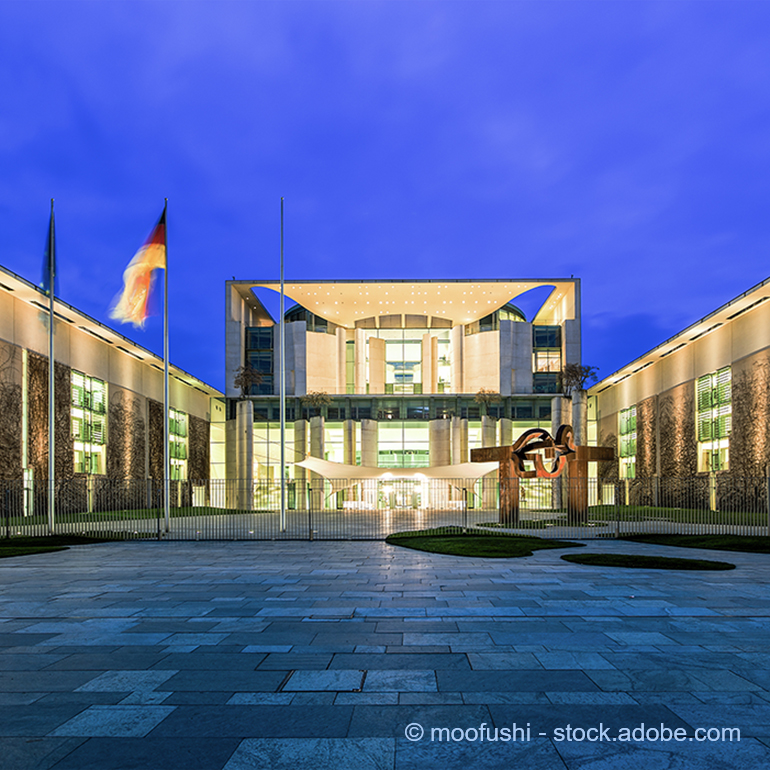The image depicts a modern government building illuminated at night, featuring a large glass facade and a distinctive roof design. Located in front is a sculpture and flagpoles, creating an inviting and contemporary public space. The sky is a deep blue, enhancing the building's features.