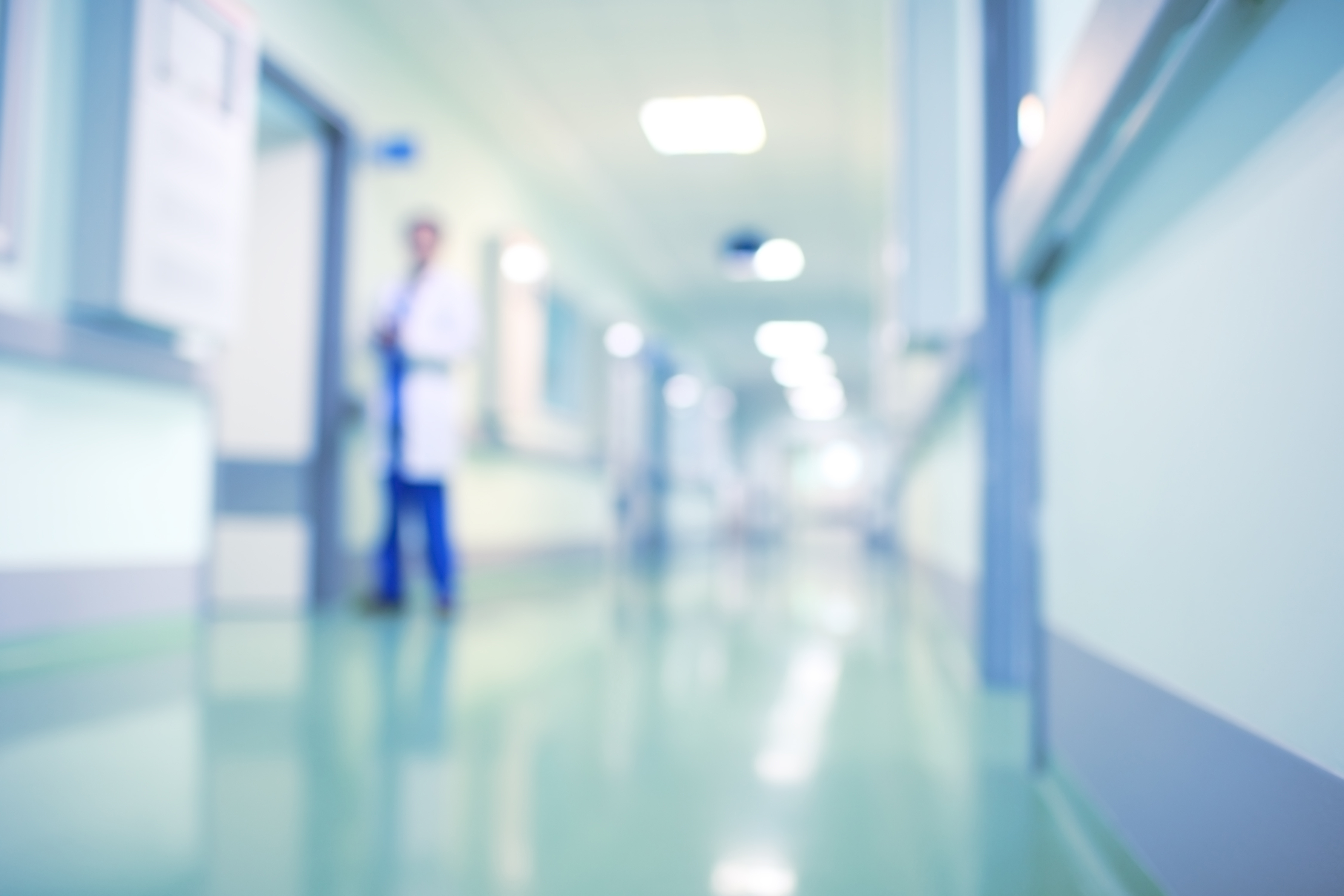 A blurred view of a hospital corridor with green flooring. A figure in a white coat and blue pants is partially visible, standing near a doorway. The setting conveys a clinical atmosphere typical of a healthcare facility.