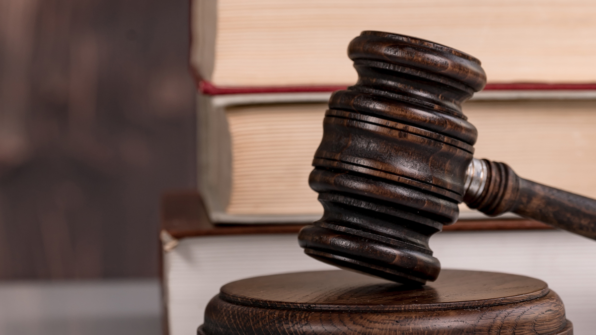 Ein Richterhammer liegt auf einem Holzblock, im Hintergrund sind zwei Bücher zu sehen. Das Bild vermittelt eine juristische Atmosphäre und symbolisiert Rechtsprechung und Gerechtigkeit.