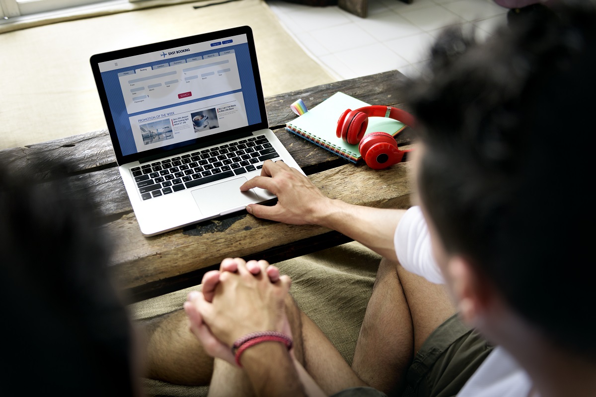 Zwei Personen sitzen an einem Holztisch und betrachten einen Laptop, der eine Webseite für Buchungen zeigt. Neben dem Laptop liegt ein Notizbuch und rote Kopfhörer. Eine Person interagiert mit dem Laptop, während die andere aufmerksam zuschaut.