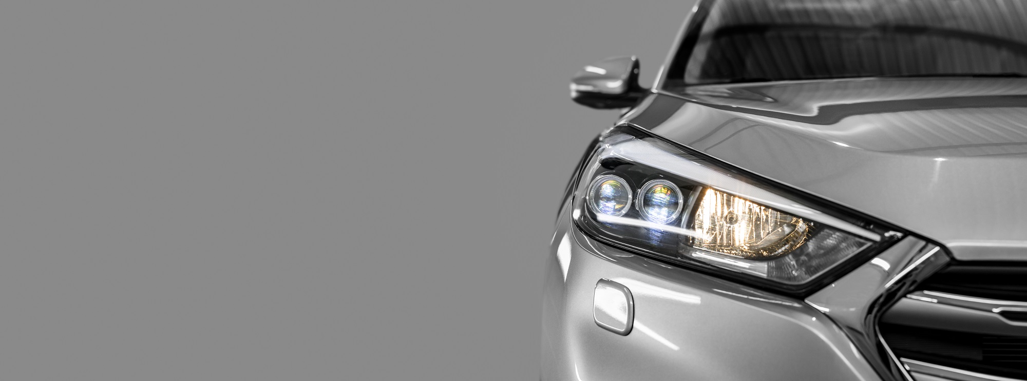 Nahaufnahme eines silbernen Autos, das die rechte Frontseite zeigt. Die Scheinwerfer sind eingeschaltet und strahlen, während der Hintergrund grau gehalten ist. Das Auto wirkt modern und elegant.