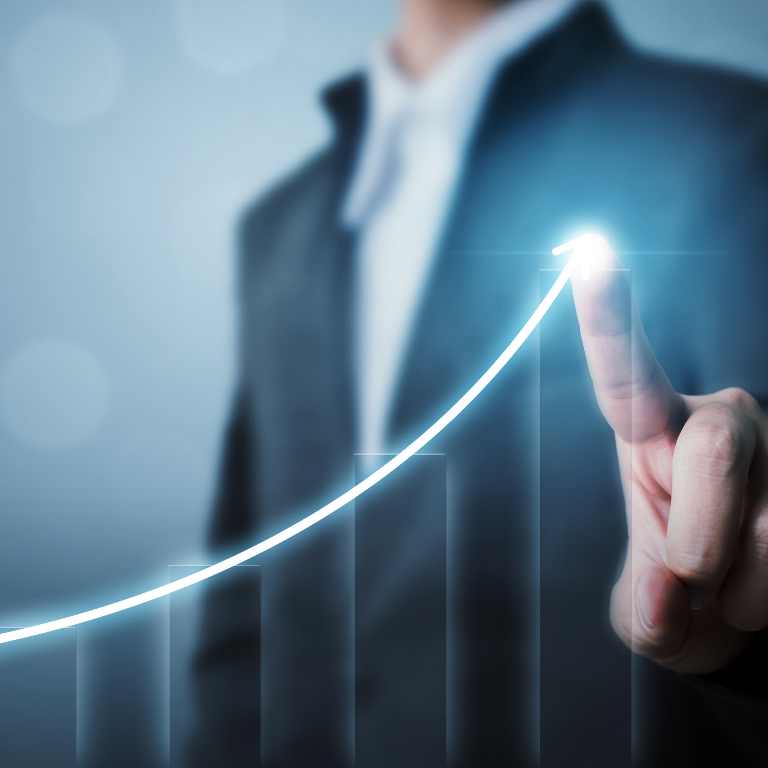Eine Person in einem Anzug zeigt mit dem Finger auf einen steigenden grafischen Verlauf, der eine positive Entwicklung symbolisiert. Im Hintergrund sind verschwommene Balkendiagramme zu erkennen. Die Gesamtstimmung vermittelt Erfolg und Wachstum.