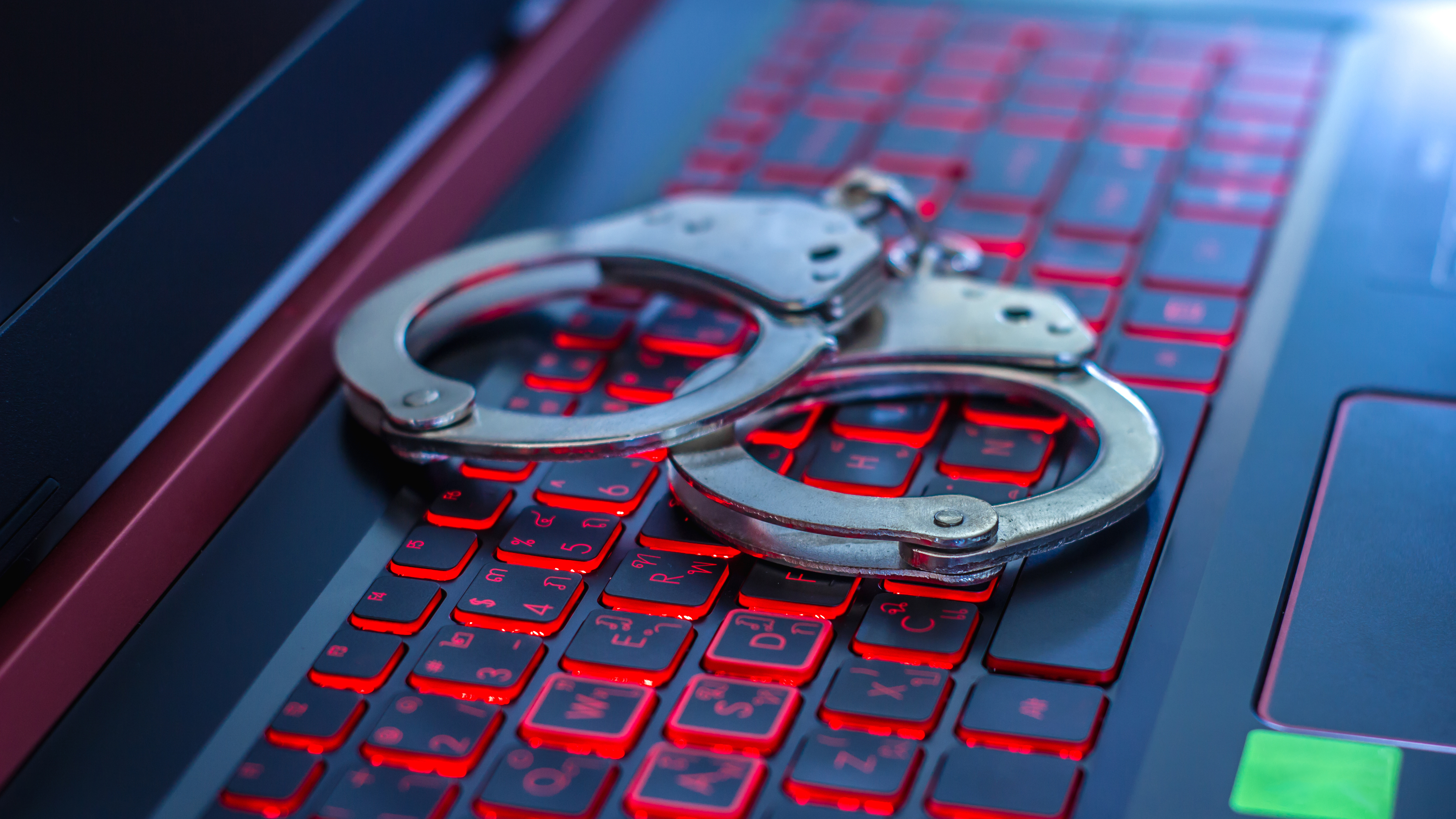 Eine Handfessel liegt auf einer beleuchteten Laptop-Tastatur. Die Tasten sind rot beleuchtet, was einen Kontrast zur dunklen Oberfläche des Laptops bildet. Die Bildkomposition vermittelt ein Gefühl von Überwachung oder Kontrolle im digitalen Raum.