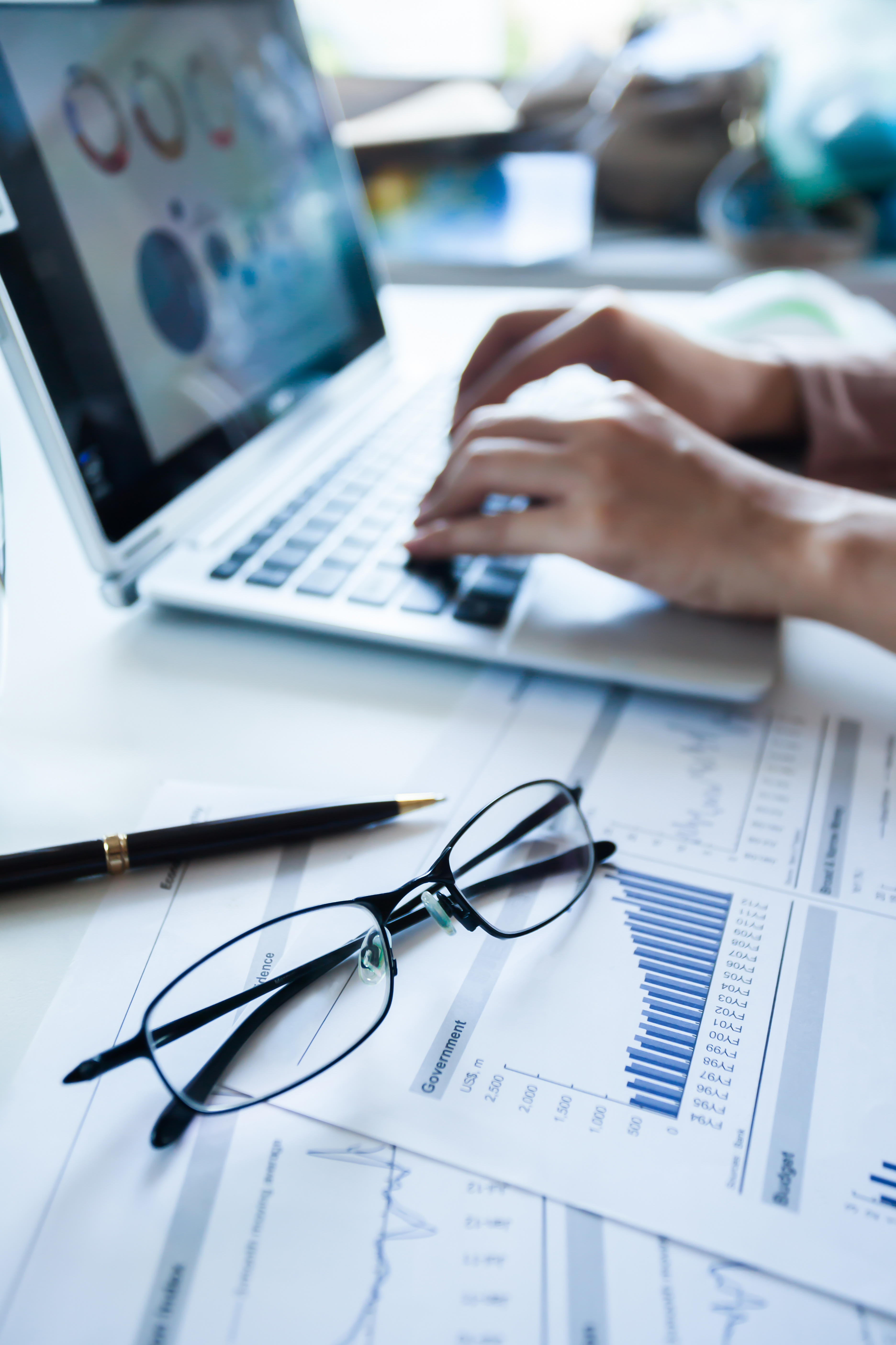A pair of black glasses rests on printed reports displaying graphs and statistics, while a person types on a laptop in the background. The scene suggests a workspace focused on data analysis and research. A pen is also visible beside the reports.
