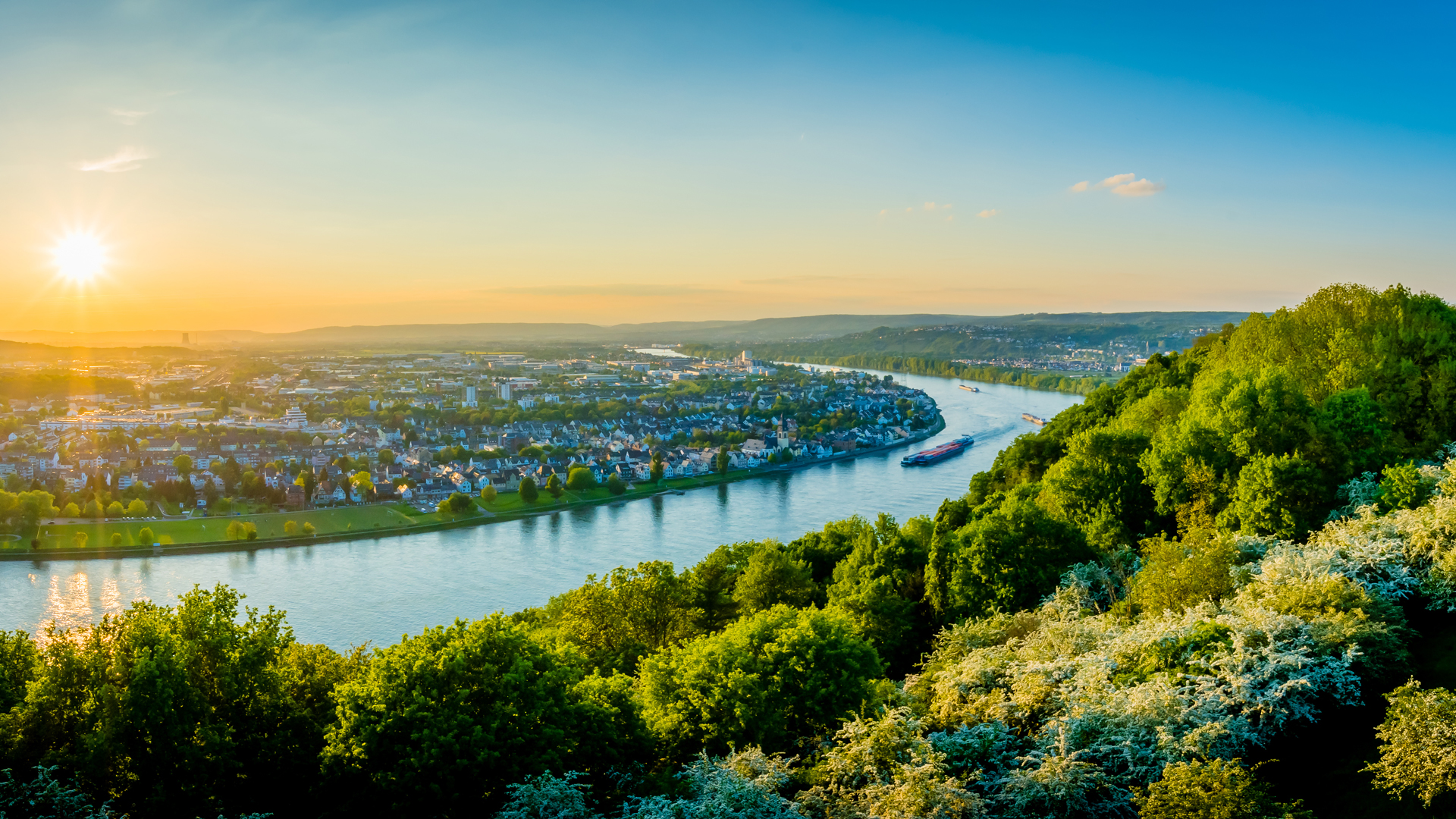 Eine malerische Aussicht auf einen Fluss, der sanft durch eine grüne Landschaft fließt. Im Hintergrund liegt eine Stadt mit Häusern, und die Sonne geht am Horizont auf, während der Himmel in warmen Farbtönen leuchtet. Ein Schiff fährt ruhig auf dem Wasser.