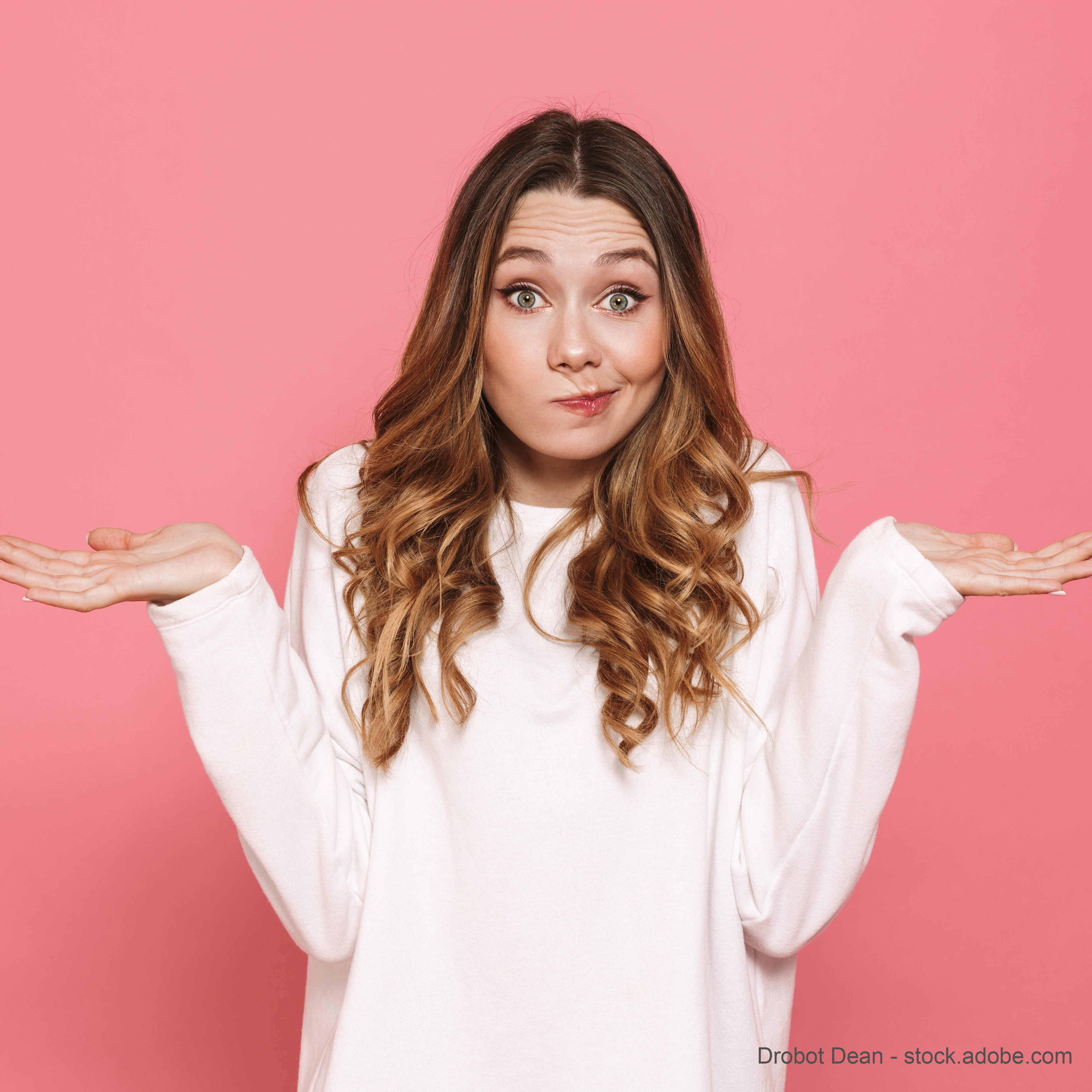 A young woman with long, wavy hair stands against a pink background. She wears a white top and makes a playful, questioning gesture by raising her hands with palms up, expressing uncertainty or confusion. Her expression is lighthearted and engaging.