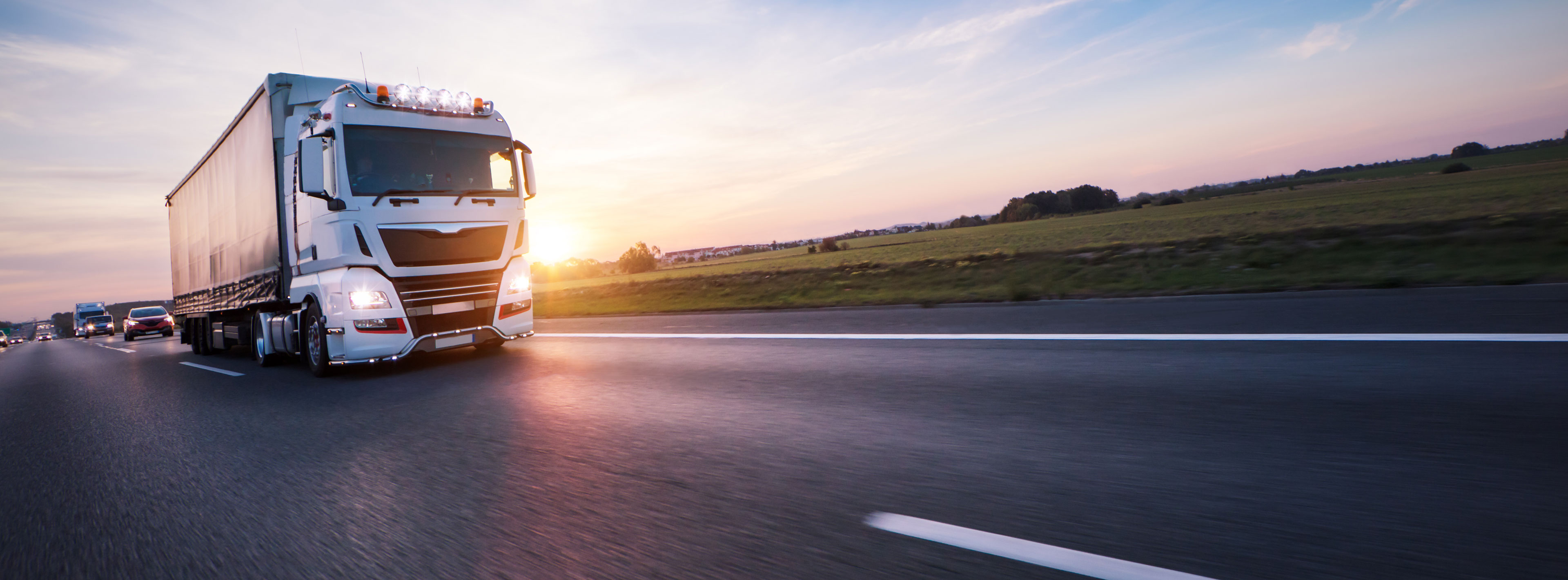 Ein großer Lastwagen fährt auf einer Landstraße, während die Sonne im Hintergrund untergeht. Die Straße ist spektral und von leichten Schatten umgeben, mit einem klaren Himmel darüber. Grüne Felder ziehen sich entlang der Straße.