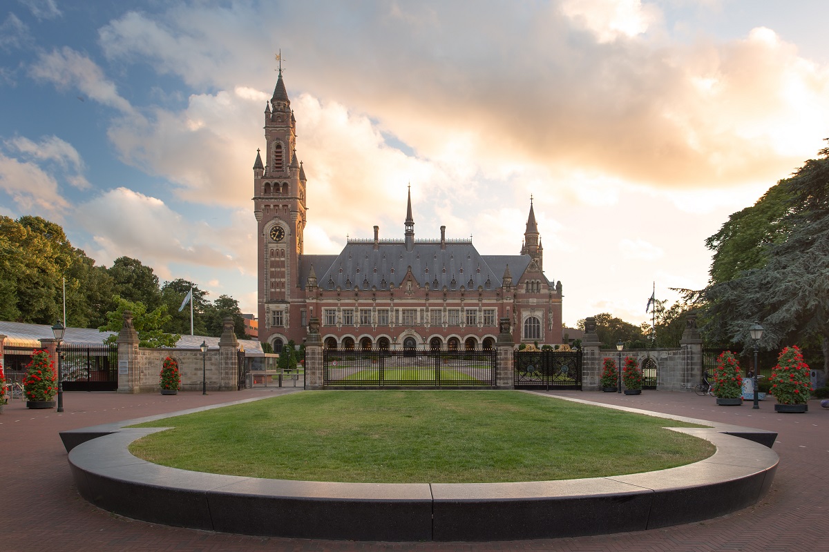 The image shows a large historical building with a tall clock tower and ornate architectural details. It is situated in a landscaped area featuring green grass and flower pots. The sky is partly cloudy, contributing to a picturesque setting.