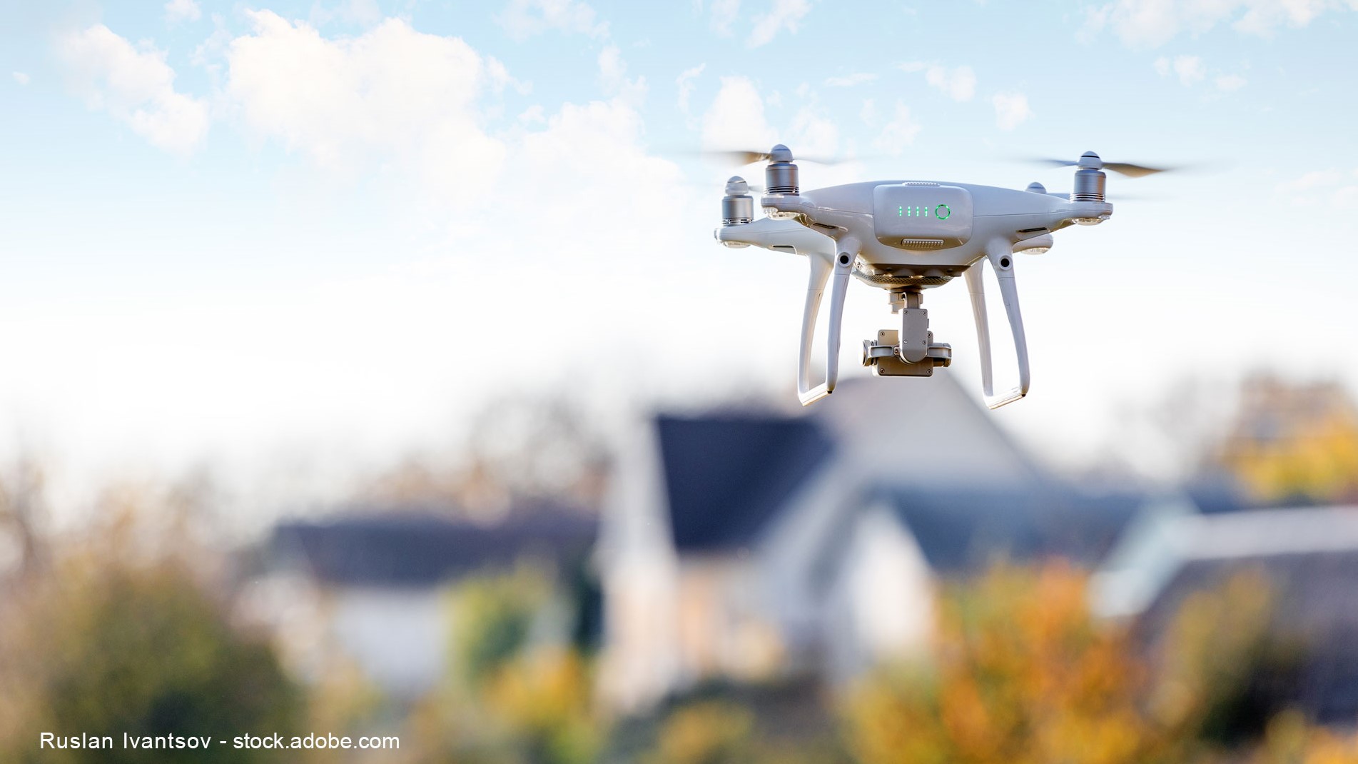 A white quadcopter drone hovers in the air against a backdrop of trees and houses. The sky is partly cloudy, creating a serene outdoor setting. The drone is equipped with a camera, suggesting it's ready for aerial photography or videography.