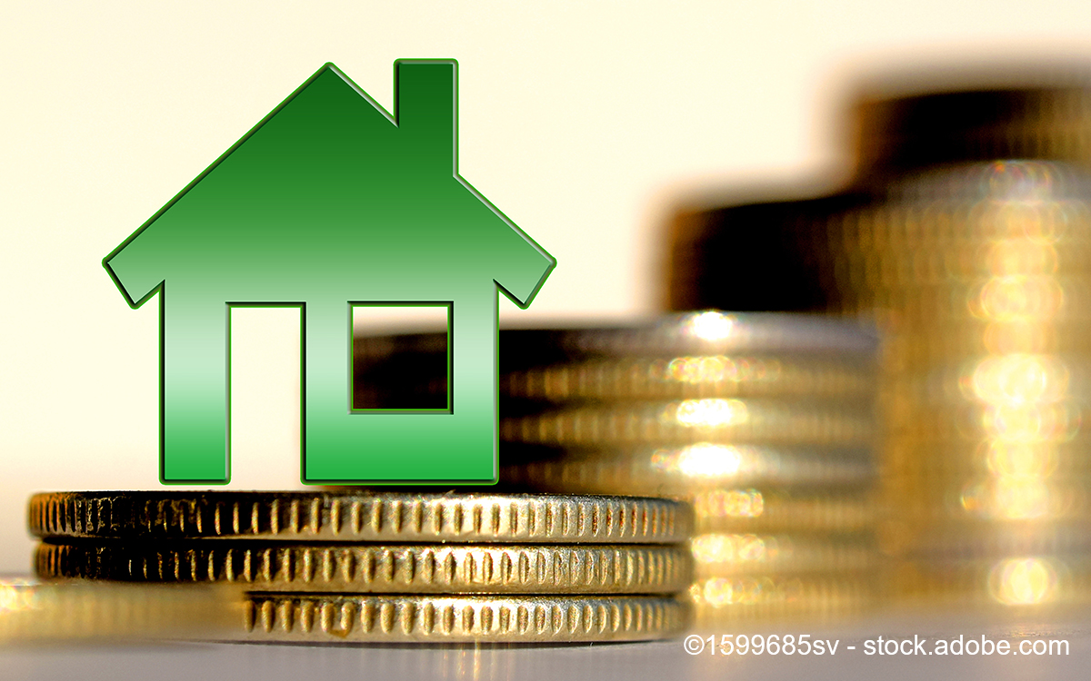 Eine grüne Hausgrafik steht im Vordergrund, vor einem unscharfen Hintergrund aus gestapelten Münzen. Die Bildkomposition vermittelt den Eindruck von Immobilieninvestitionen oder finanzieller Sicherheit.
