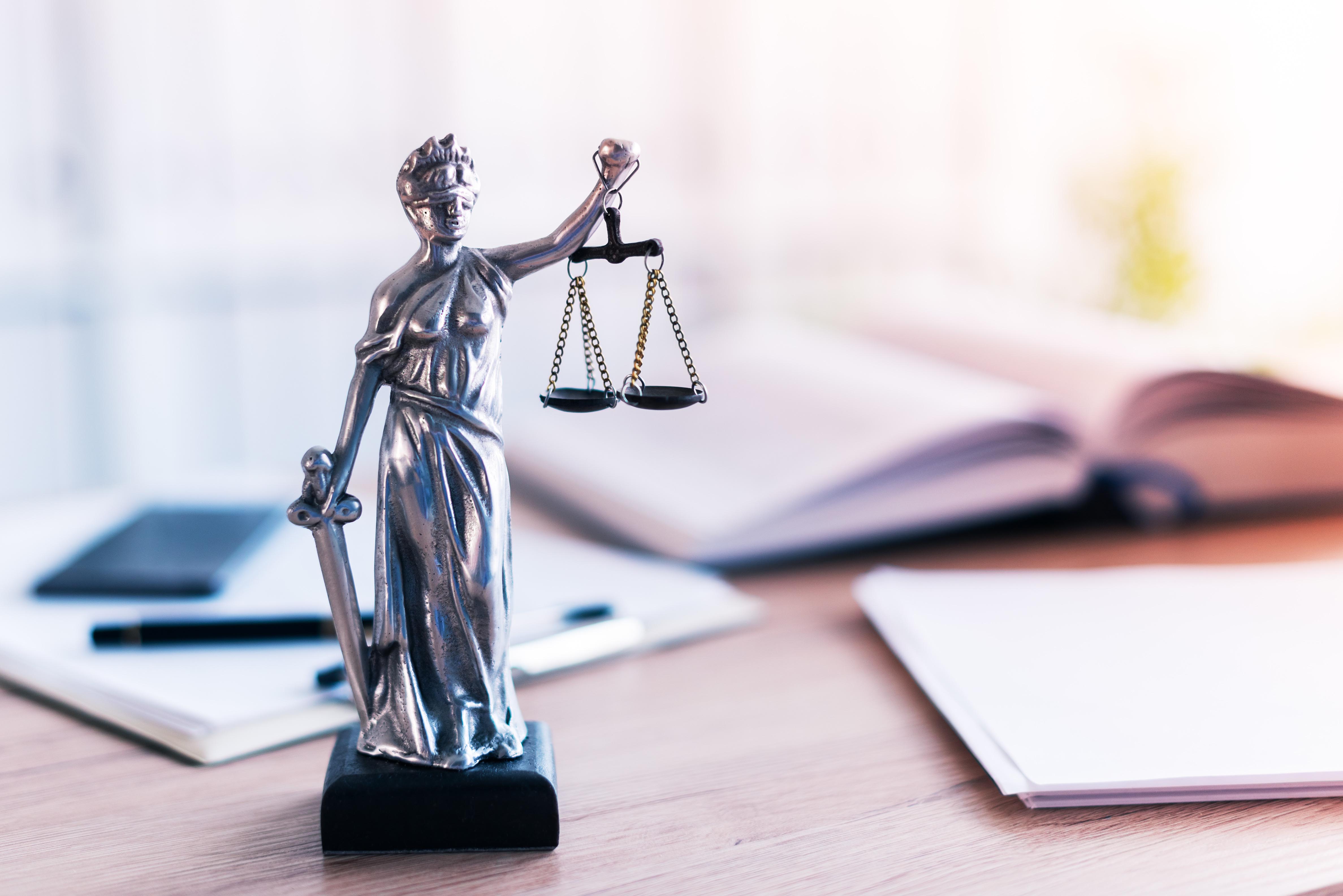 A silver statue of Lady Justice stands holding a set of balanced scales in one hand and a sword in the other. The background features blurred legal books and documents, representing themes of law, justice, and legal proceedings.