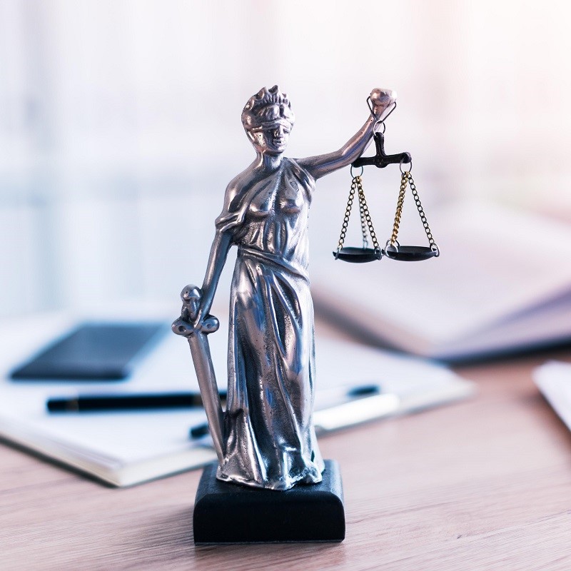 Eine metallene Statue der Justitia, die mit einer Hand eine Waage und mit der anderen ein Schwert hält. Die Figur symbolisiert Gerechtigkeit und Rechtsprechung. Der Hintergrund zeigt unscharfe Büromaterialien.