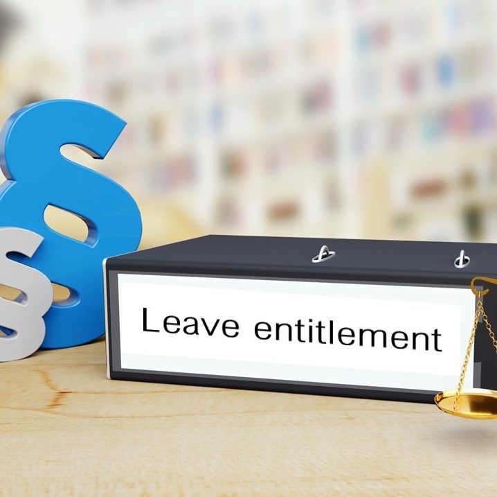 A binder labeled "Leave entitlement" sits on a desk, accompanied by large decorative legal symbols. A small scale representing justice is attached to the binder, emphasizing a legal or regulatory theme related to employee leave rights.