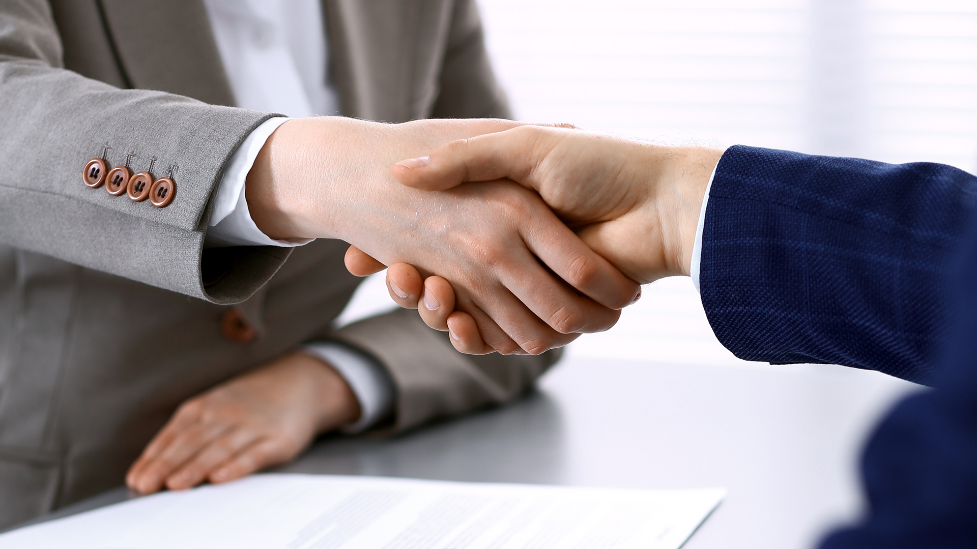 Zwei Personen schütteln sich die Hände, was einen Vertrag oder eine Vereinbarung symbolisiert. Die Hände sind gut sichtbar, und die Umgebung wirkt professionell. Im Hintergrund sind Dokumente auf einem Tisch zu erkennen, was auf eine geschäftliche Situation hinweist.