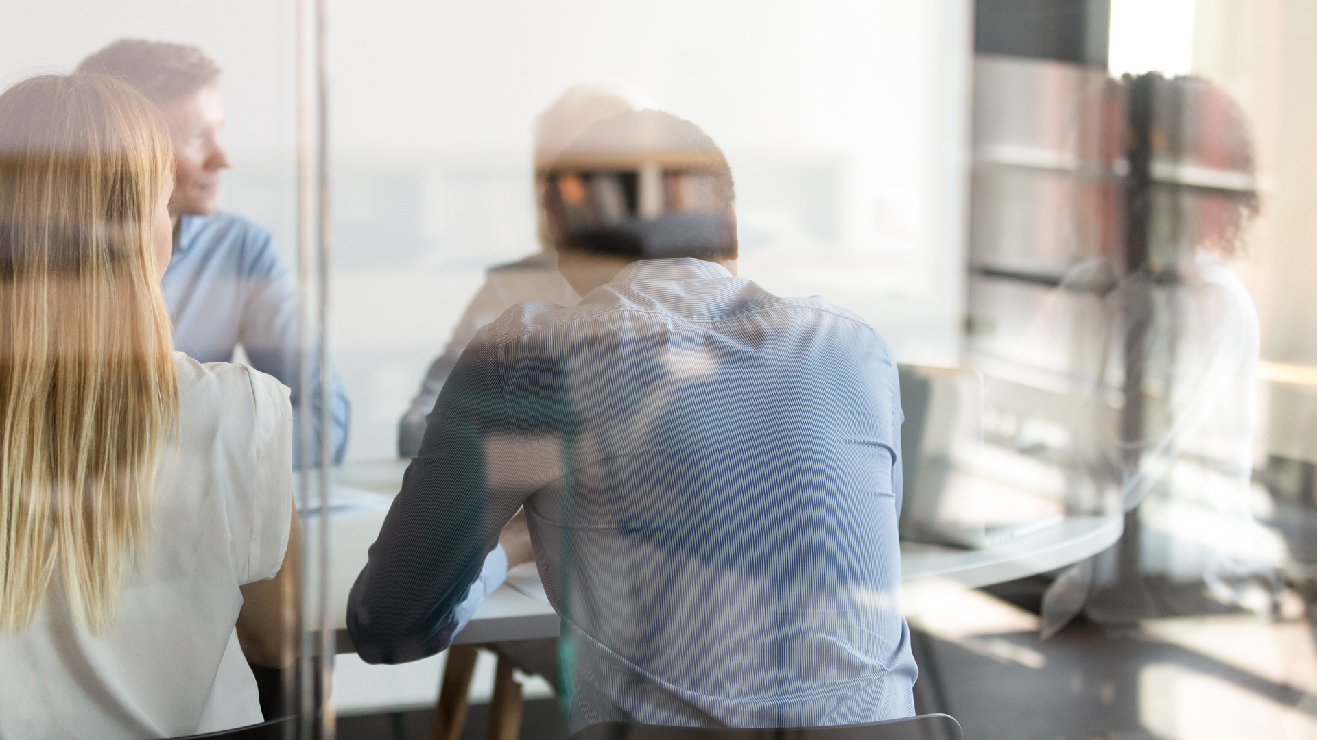 Eine Gruppe von Personen sitzt an einem Tisch in einem Besprechungsraum. Man sieht die Rückseiten von mehreren Köpfen. Die Atmosphäre wirkt professionell und konzentriert, während sie miteinander diskutieren. Licht strömt durch große Fenster und schafft eine helle Umgebung.