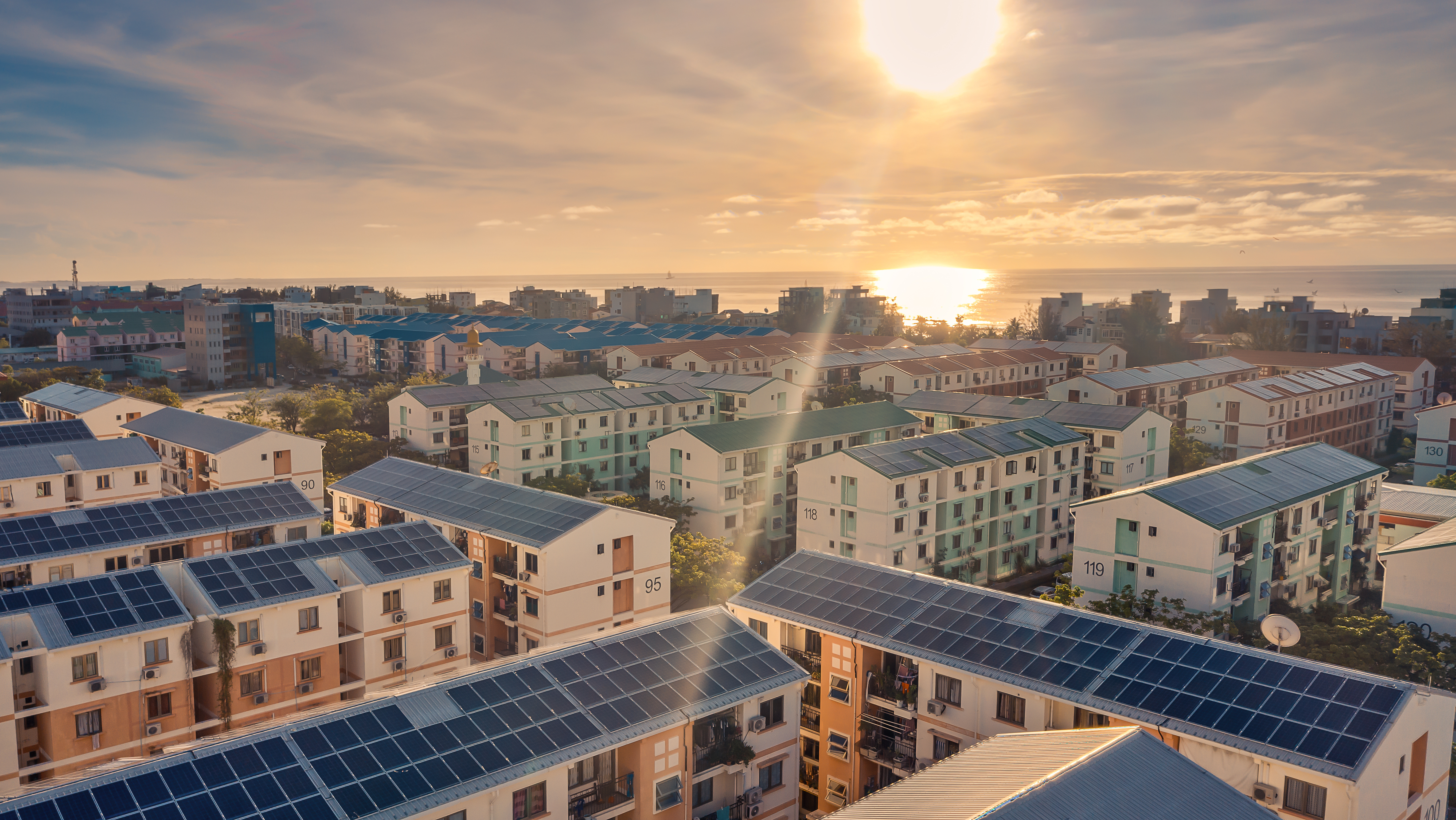 Luftaufnahme einer Wohnsiedlung mit mehreren Gebäuden, die mit Solarpanelen auf den Dächern ausgestattet sind. Im Hintergrund sind weitere Gebäude und das Meer bei Sonnenuntergang zu sehen, was eine friedliche Atmosphäre vermittelt.