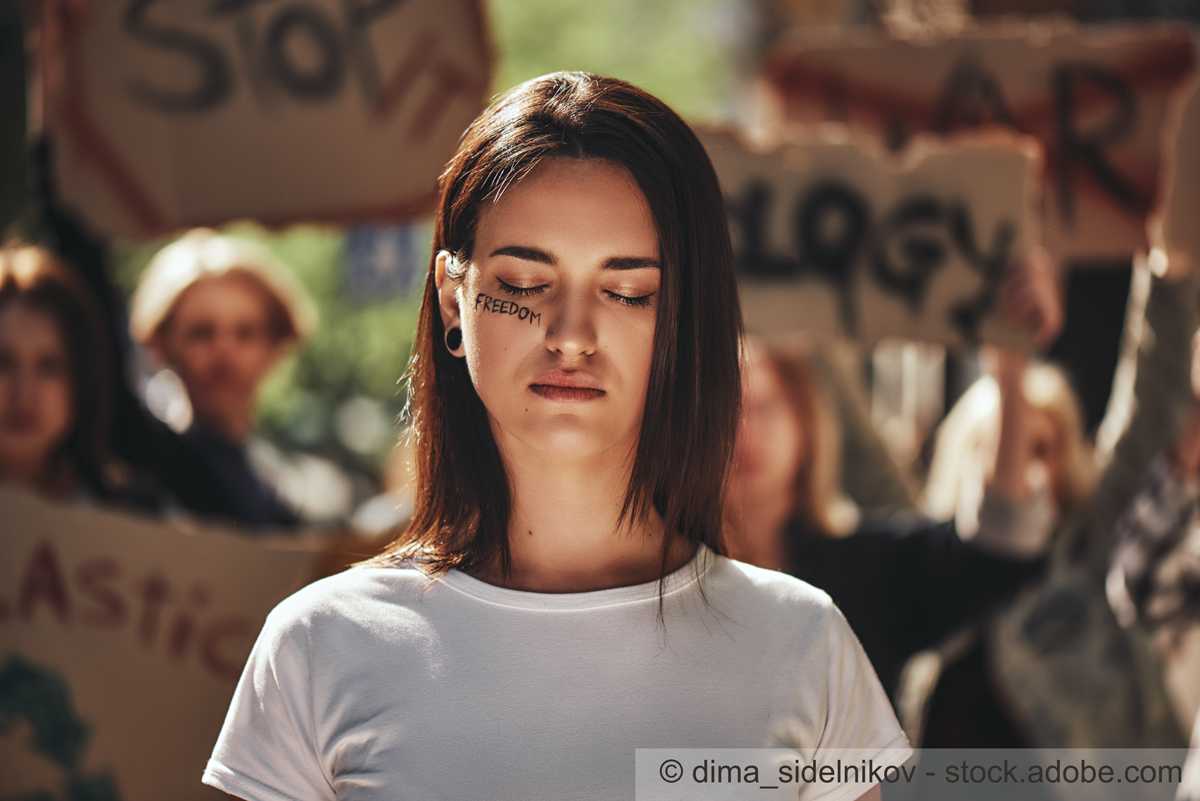 A young woman with a solemn expression stands with her eyes closed, wearing a white shirt. The word "FREEDOM" is written on her face. In the background, people hold up protest signs, emphasizing a message of activism and social justice.