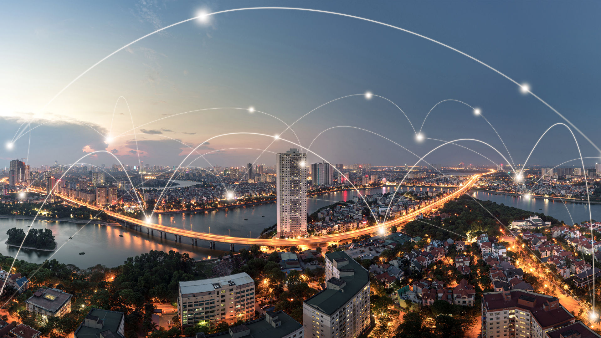 Eine Stadtansicht bei Dämmerung zeigt ein lebhaftes urbanes Gebiet mit einer großen Brücke über einen Fluss. Über der Szenerie verlaufen lichtartige Linien, die Netzwerke oder Verbindungen symbolisieren, was das Thema Technologie und Vernetzung verdeutlicht.