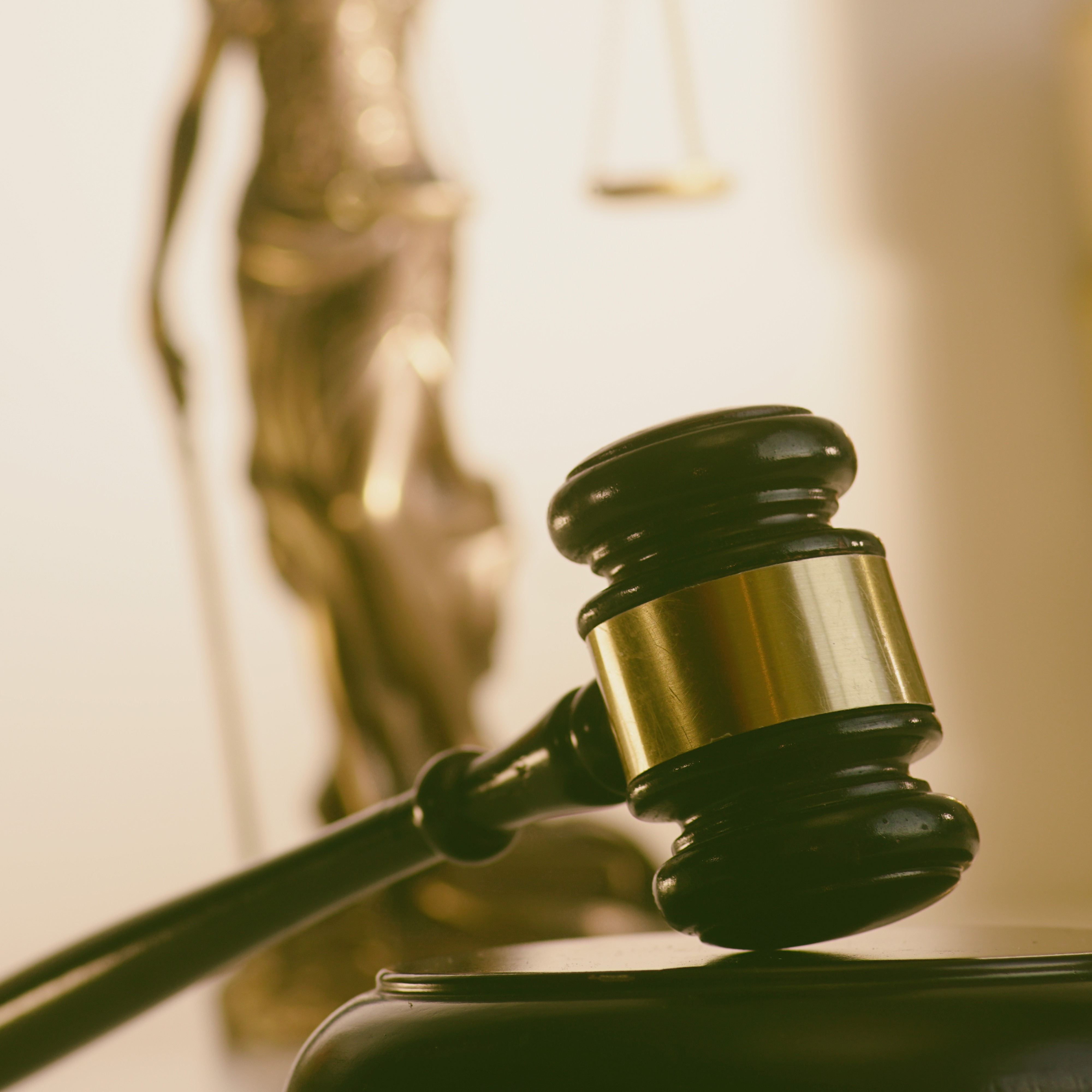 A gavel rests on a block, symbolizing justice and the legal system. In the background, a statue representing justice stands, holding scales, emphasizing the theme of law and fairness. The image conveys authority and the courtroom environment.
