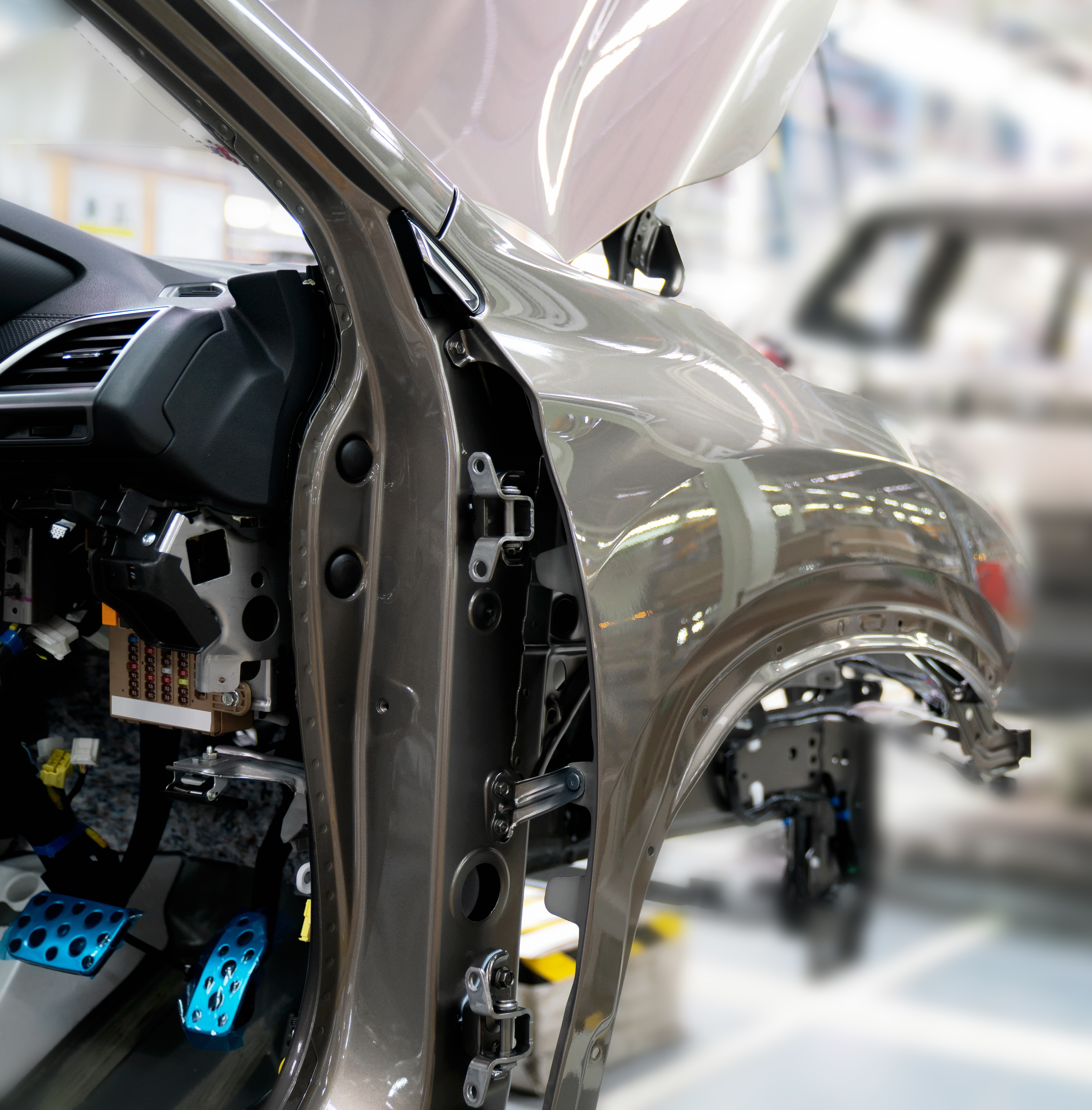 Eine Nahaufnahme eines Autoinnenraums während der Montage. Man sieht die offene Fahrzeugtür mit sichtbaren Komponenten wie Pedalen und Anbauteilen. Der Hintergrund zeigt eine Produktionsumgebung mit verschwommenen Fahrzeugen und Werkzeugen.
