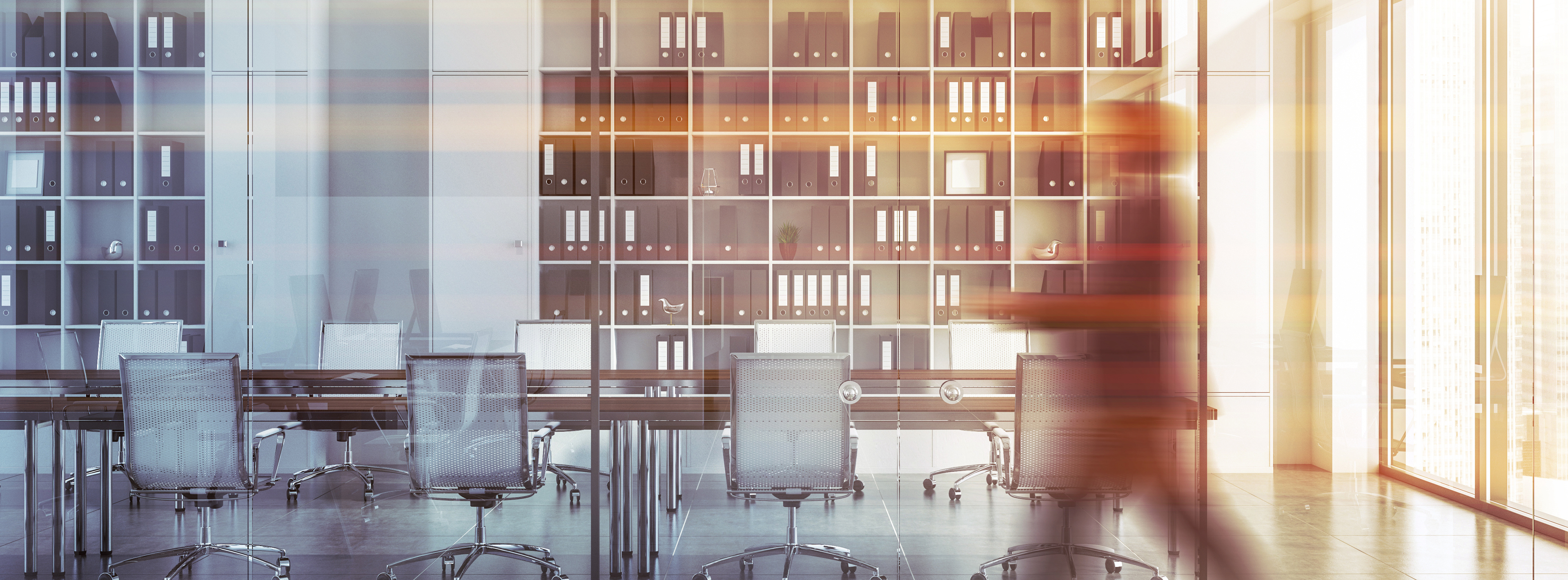 A modern office space features a long conference table surrounded by transparent chairs. Behind the table is a shelf filled with neatly organized black folders. A person walks past in a blur, suggesting an active work environment with ample natural light.