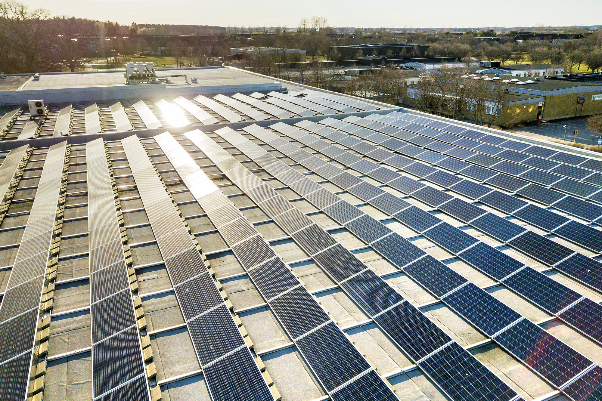 Aerial view of a large commercial roof covered with solar panels, reflecting sunlight. The panels are arranged in neat rows, showcasing a focus on renewable energy. In the background, other rooftops and greenery are visible, emphasizing an urban setting.