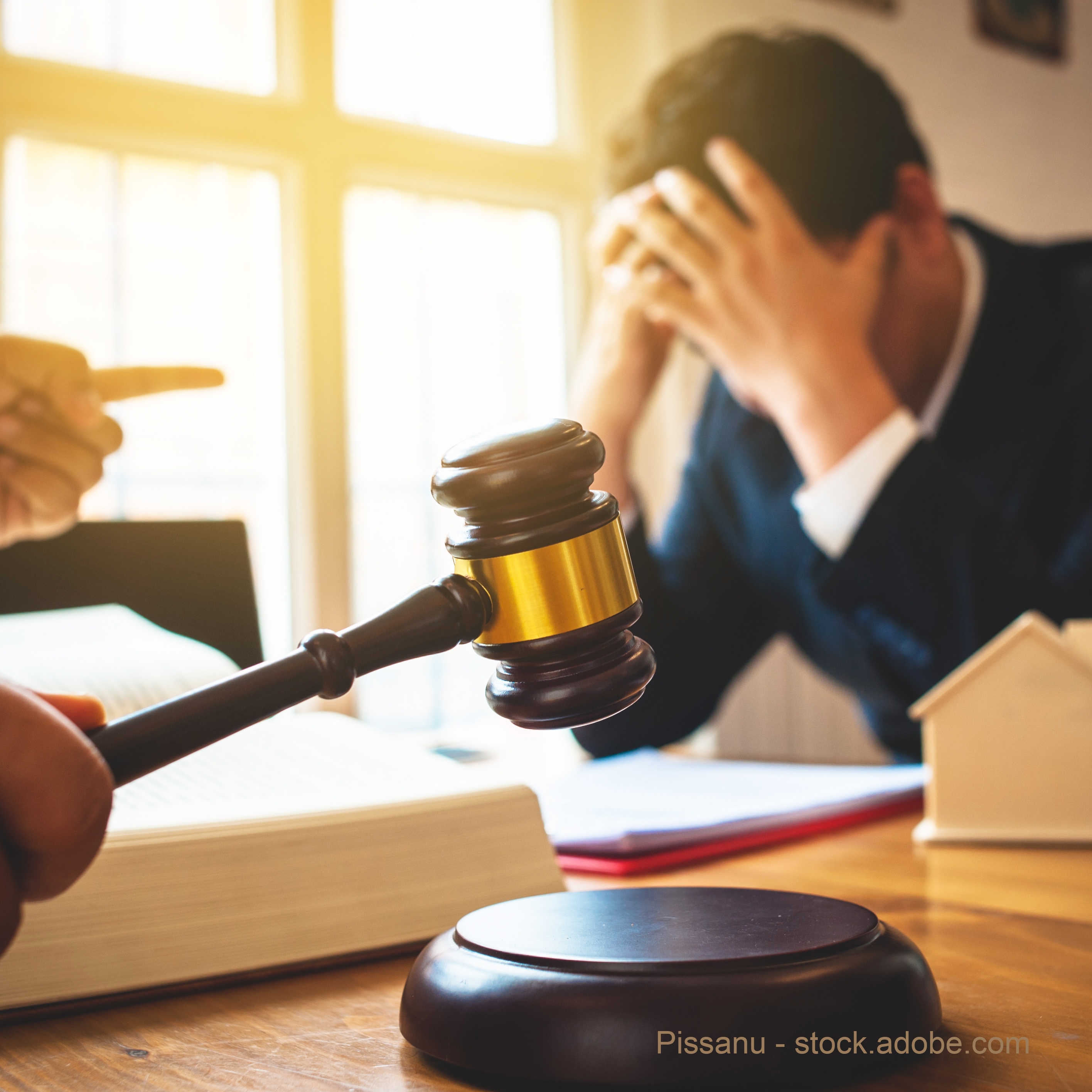 A gavel is raised in the foreground, symbolizing authority, while a distressed man with his head in his hands sits at a table covered with legal documents. Another person is pointing, suggesting a confrontation or disagreement. The scene conveys tension related to a legal matter.