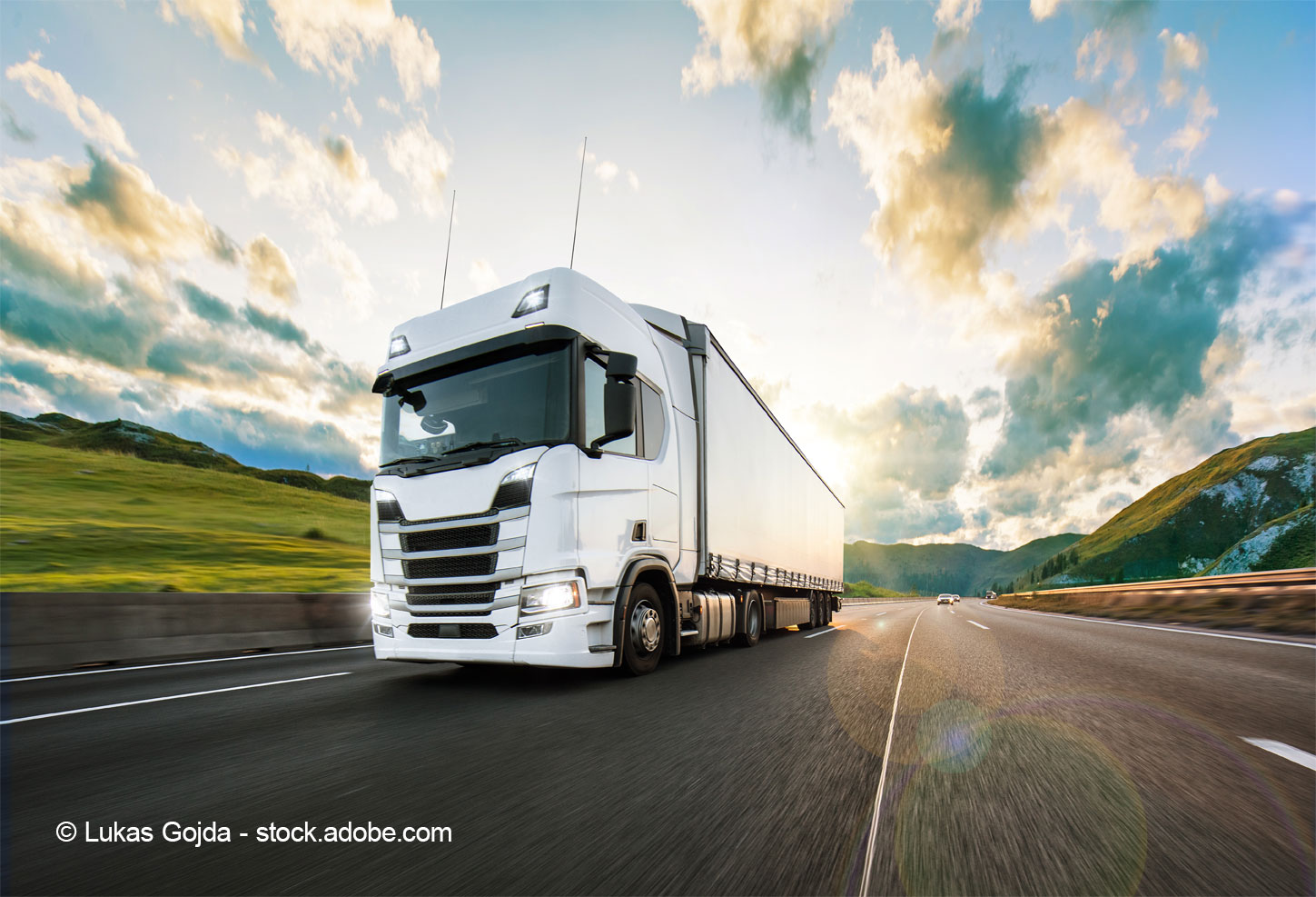 A large white truck is driving on a highway, showcasing a scenic landscape with rolling hills and a cloudy sky in the background. The truck is in motion, conveying a sense of travel and transportation.