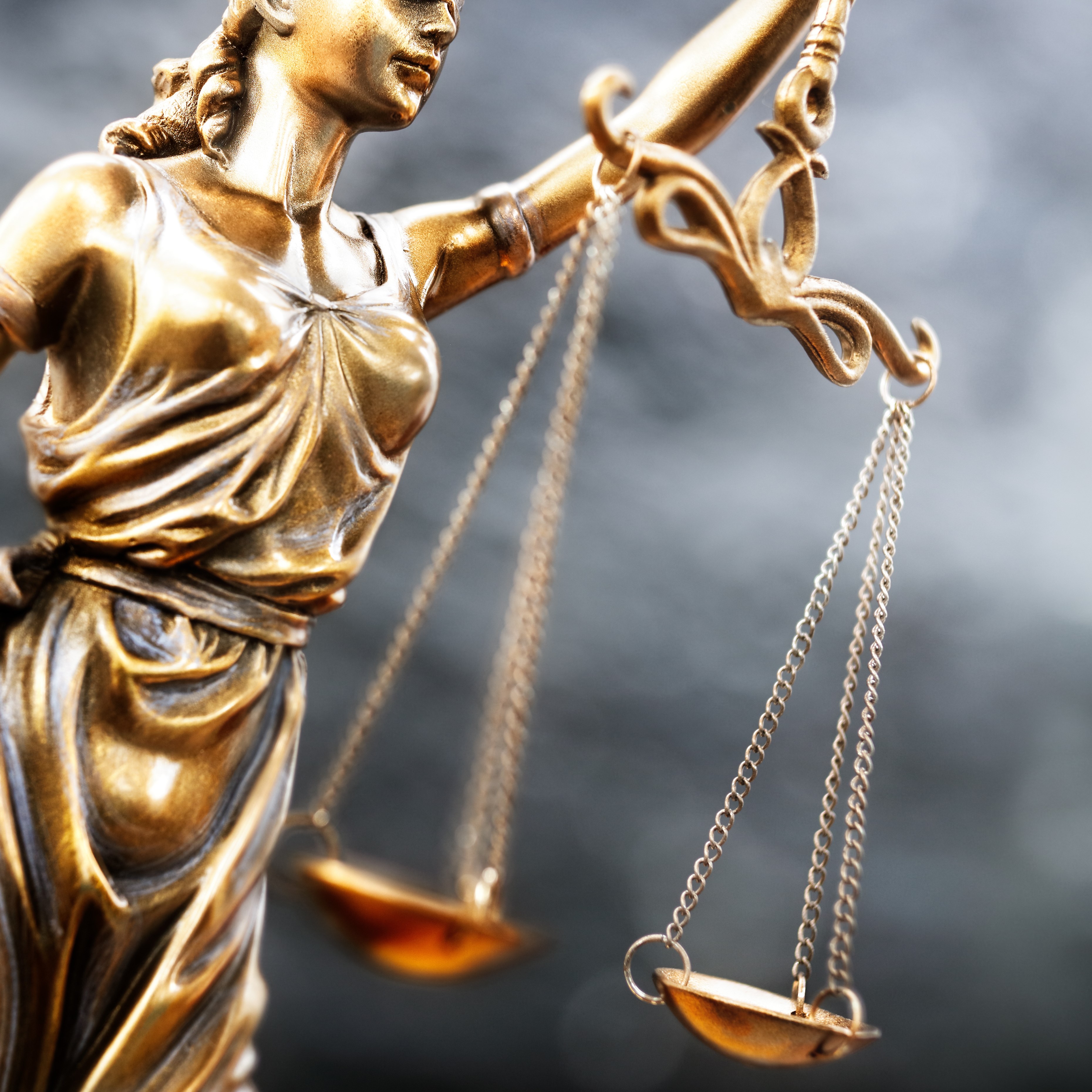 A close-up view of a golden statue representing Lady Justice. She holds a set of balancing scales in her right hand, symbolizing fairness and the law. Her expression is serene, reflecting impartiality, while she stands draped in flowing robes. The background is softly blurred.