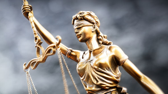 Eine goldene Statue von Justitia, blind mit verbundenen Augen, hält in der rechten Hand eine Waage und in der linken ein Schwert. Sie symbolisiert Gerechtigkeit und Unparteilichkeit. Der Hintergrund ist unscharf, um den Fokus auf die Statue zu legen.