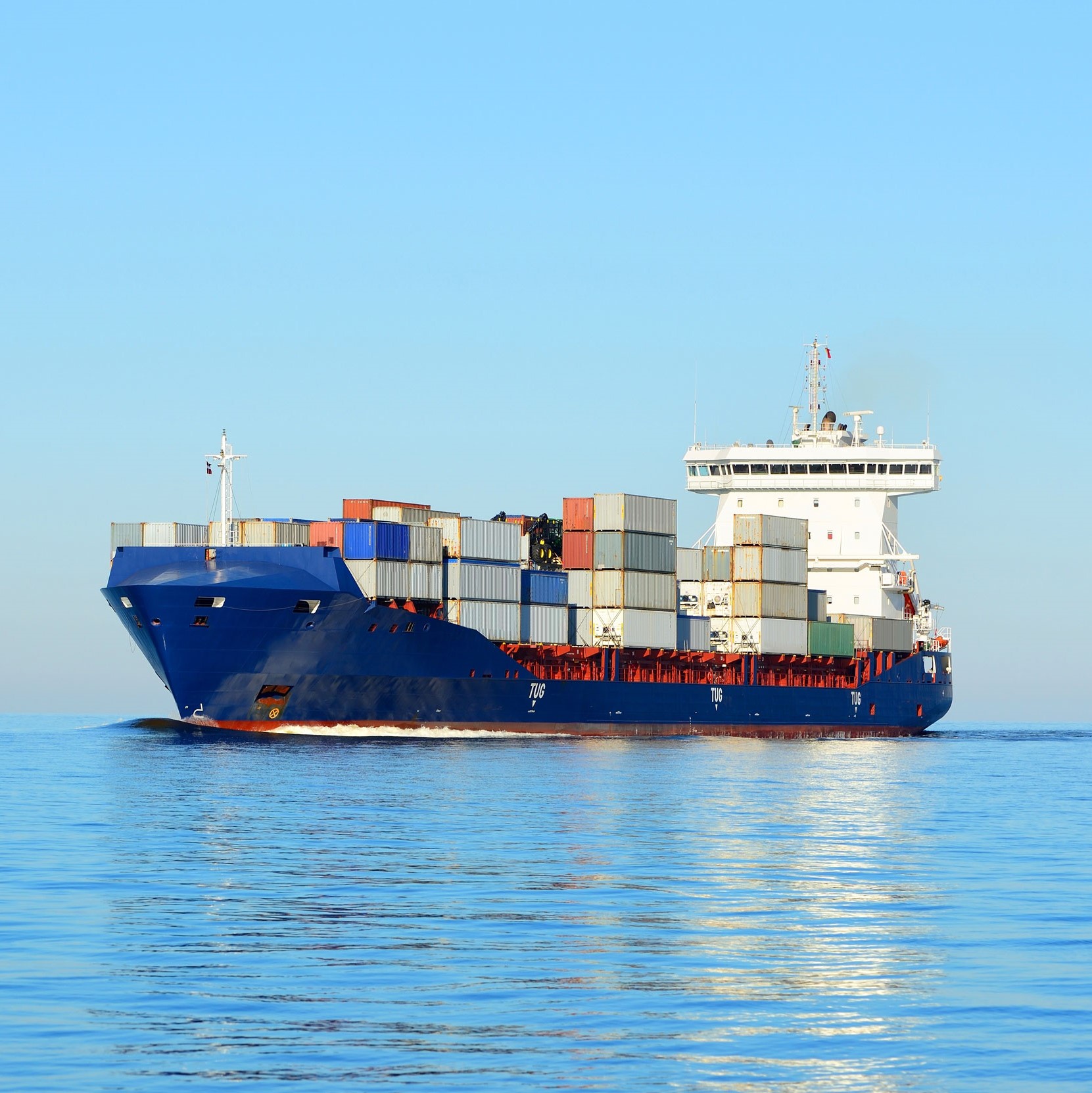 Ein großes Containerschiff mit zahlreichen bunten Containern fährt über ruhiges Wasser. Der Himmel ist klar und blau, was eine friedliche maritime Szene schafft.