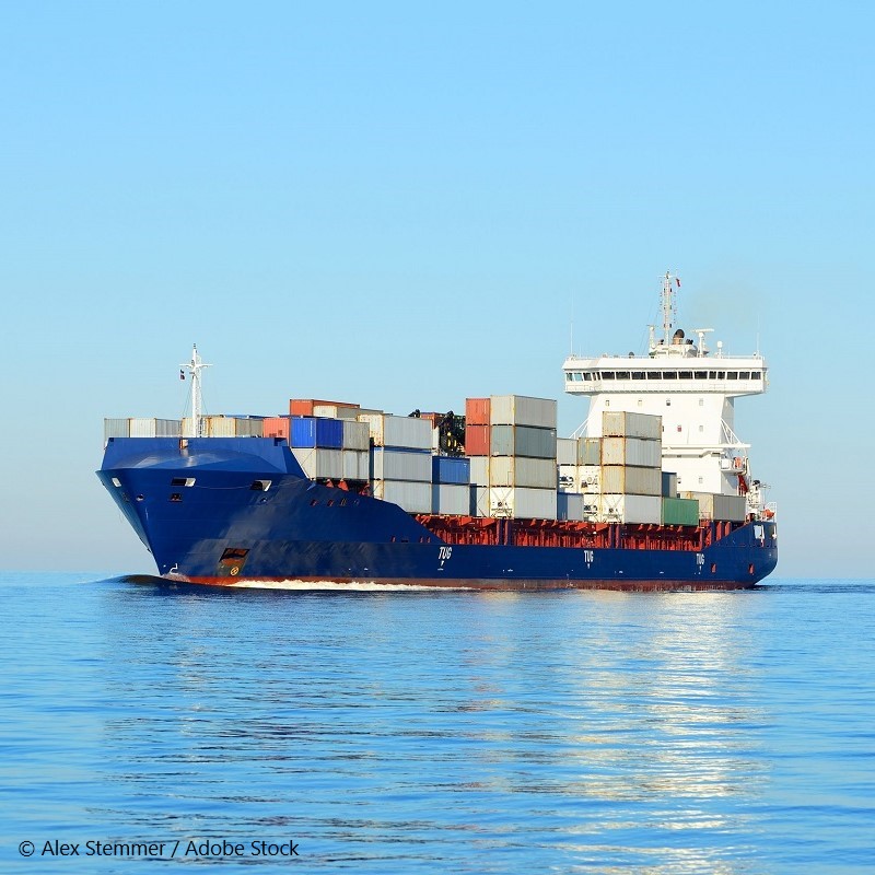 A large container ship sails on calm waters, carrying various stacked cargo containers. The sky is clear, and the ship appears sturdy and well-maintained, indicating its role in maritime transport.