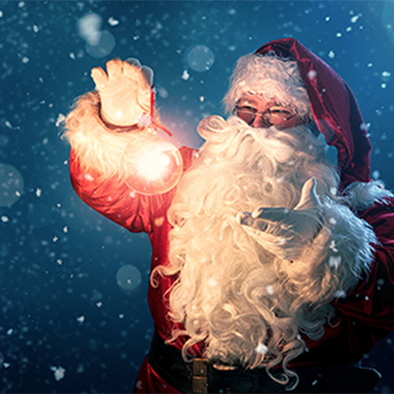 A cheerful Santa Claus in a red suit and hat holds a glowing ornament in one hand while gesturing with the other. Snowflakes fall around him against a dark blue background, creating a festive holiday atmosphere.