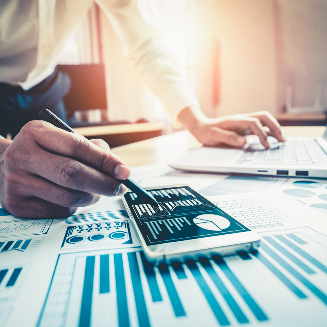 Eine Hand hält einen Stift und tippt auf einem Smartphone, das Datenvisualisierungen zeigt. Im Hintergrund steht ein Laptop, während auf einem Tisch Diagramme und Tabellen liegen. Die Szene vermittelt ein Gefühl von Produktivität und Datenanalyse im Arbeitsumfeld.