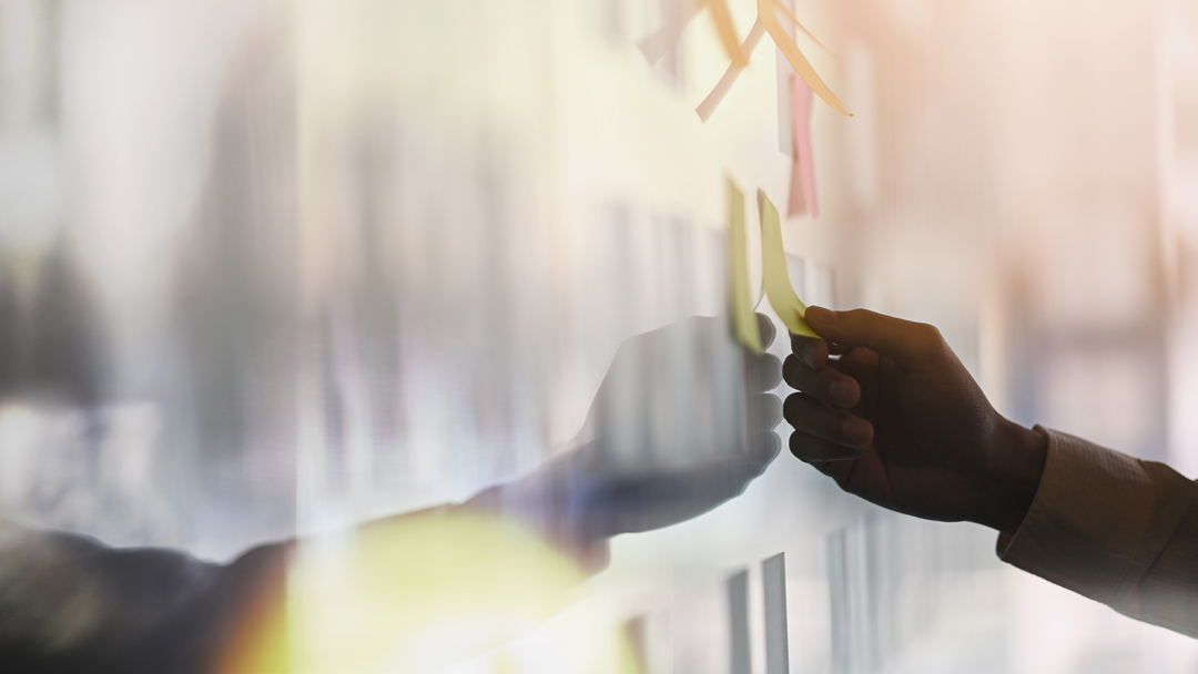 Eine Hand befestigt einen bunten Haftnotizzettel an einer Wand. Die Wand ist mit mehreren Haftnotizen in verschiedenen Farben dekoriert, die Informationen oder Ideen darstellen könnten. Das Bild vermittelt einen Eindruck von Kreativität und Teamarbeit.