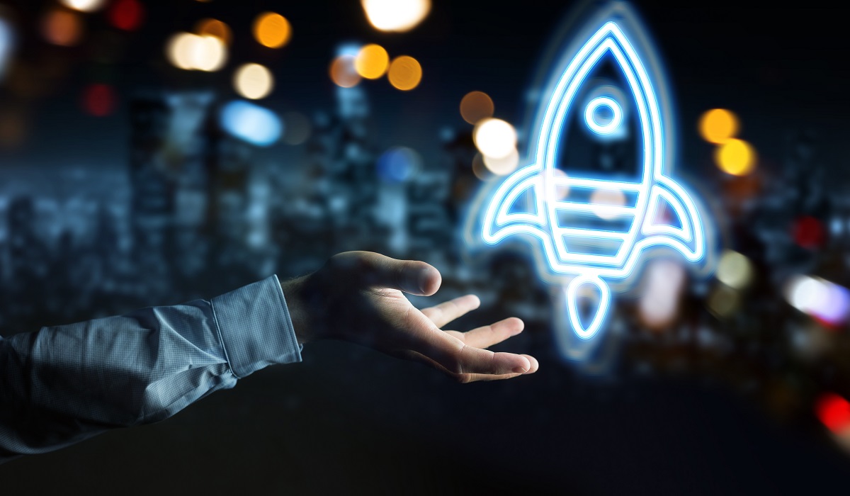 A hand reaching out to display a glowing illustration of a rocket. The background features blurred city lights, creating a dynamic and futuristic atmosphere. The image symbolizes innovation, potential, and aspiration.