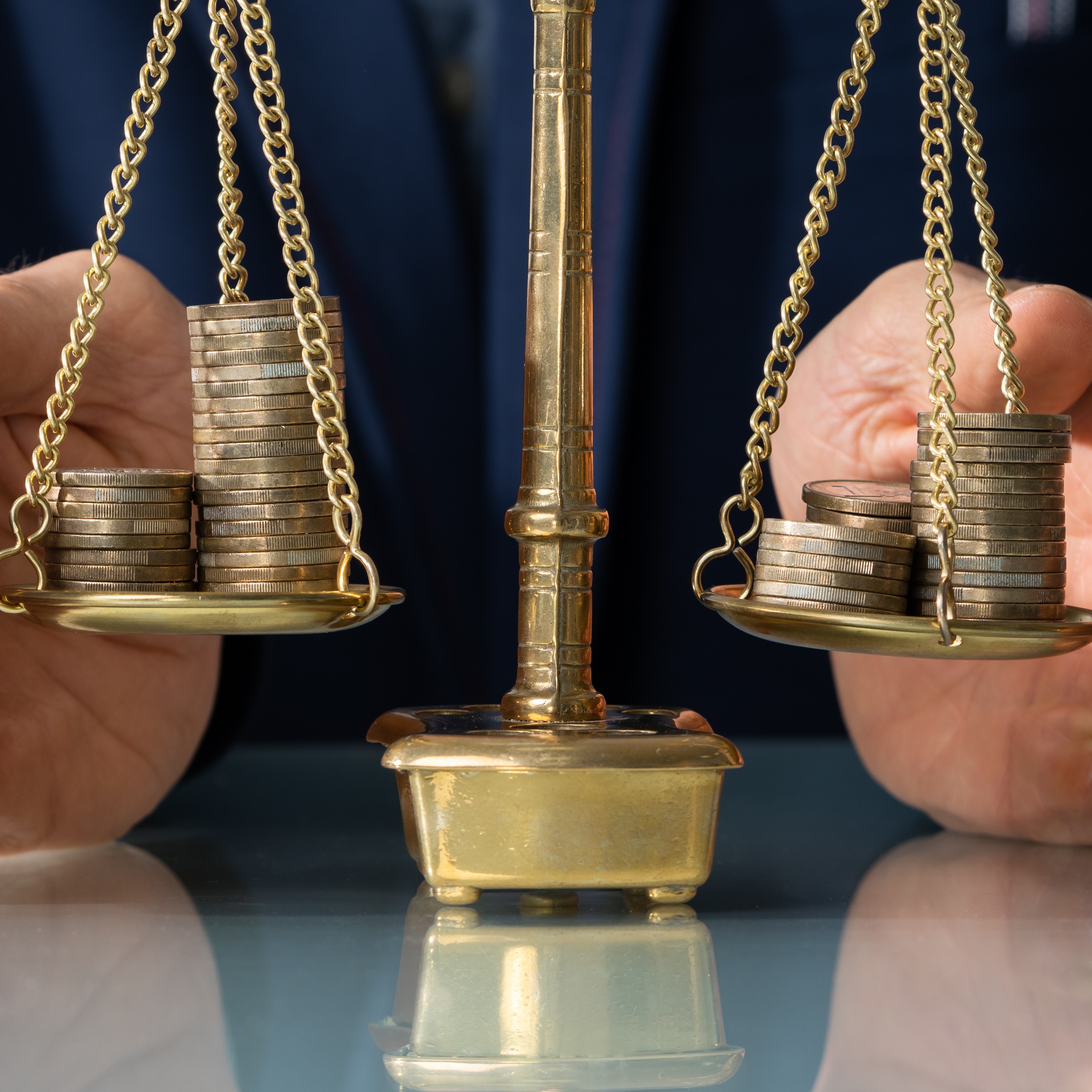 Eine Waage steht in der Mitte, auf beiden Seiten sind kleine Stapel von Münzen abgebildet. Die Waage symbolisiert den Ausgleich oder die Balance zwischen Werten. Hände halten die Waage, was auf einen beabsichtigten Vergleich hindeutet.