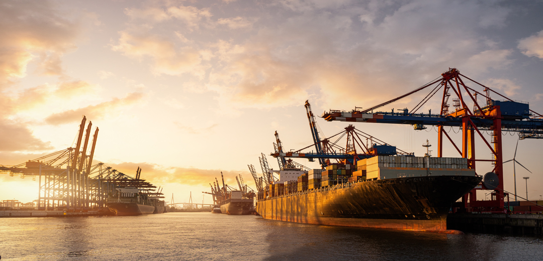 Ein Hafen bei Sonnenuntergang mit mehreren großen Containerschiffen im Wasser. Kräne heben Container von den Schiffen. Die Szene strahlt eine ruhige, industrielle Atmosphäre aus, während die letzten Sonnenstrahlen die Schiffe beleuchten.