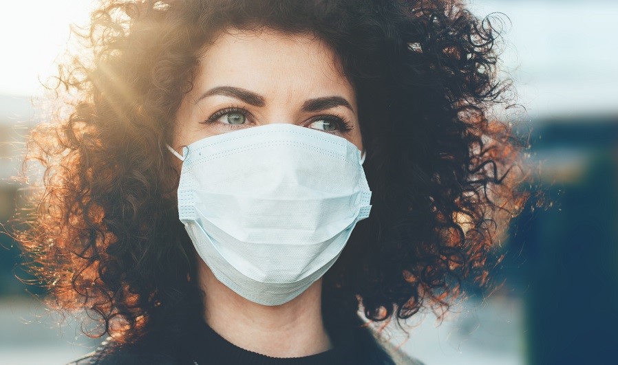A woman with curly hair is wearing a blue surgical mask. She is looking slightly off-camera, with sunlight creating a warm glow around her. The focus is on her eyes, which express a sense of determination and awareness.
