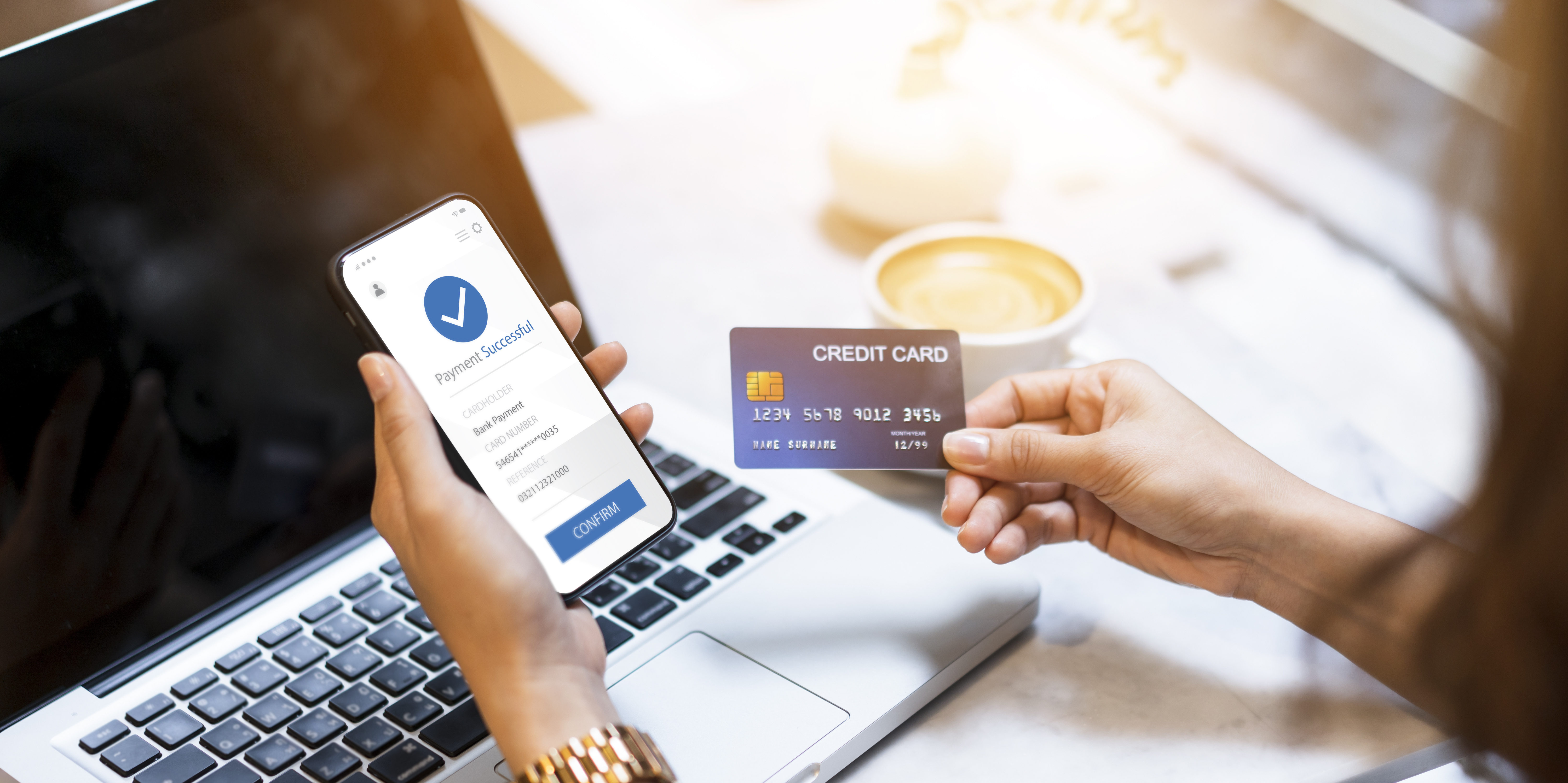 Eine Person hält ein Smartphone mit der Bestätigung einer erfolgreichen Zahlung in der einen Hand und eine Kreditkarte in der anderen. Im Hintergrund ist ein Laptop zu sehen, der auf einem Tisch steht.