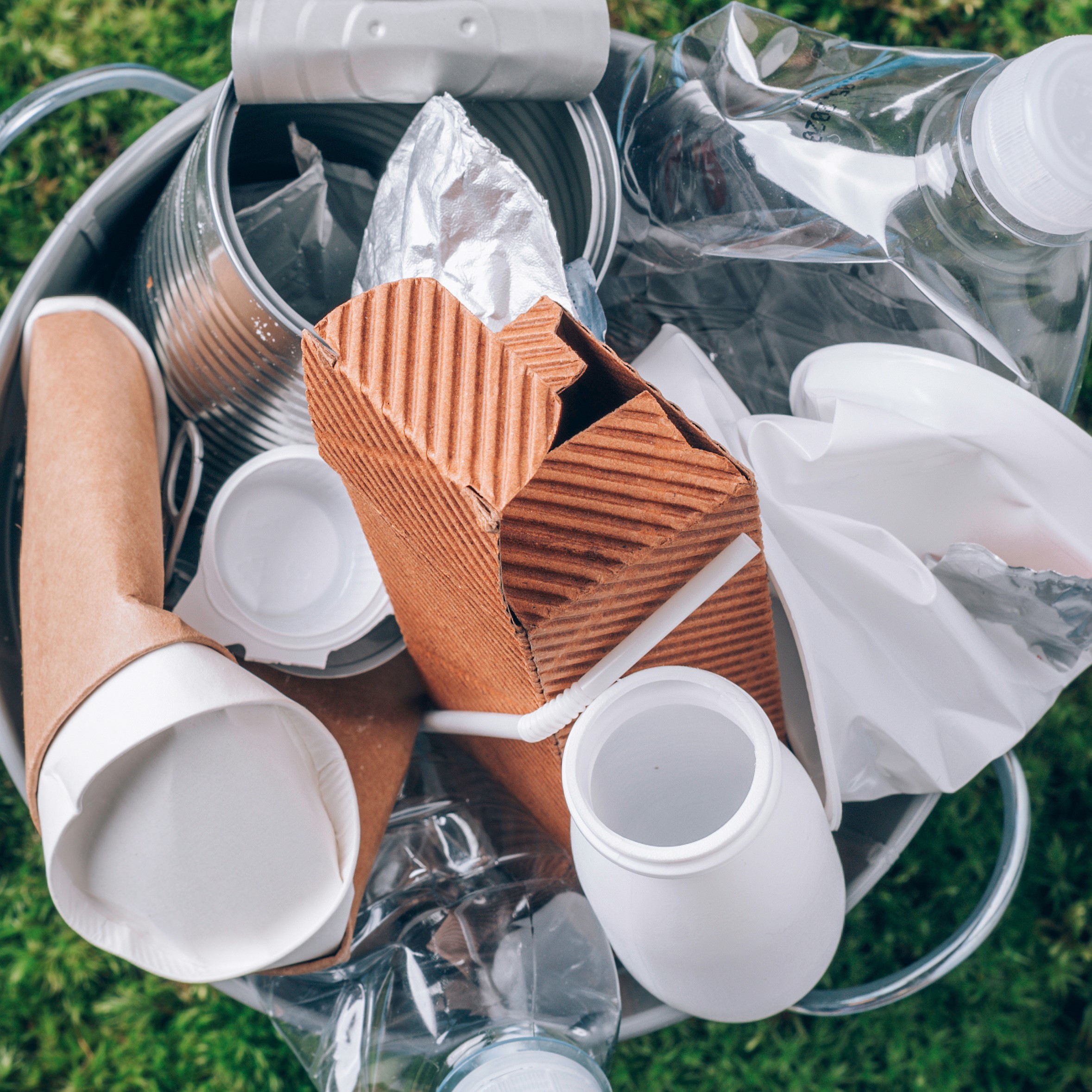 Der Bildinhalt zeigt einen überfüllten Abfalleimer mit verschiedenen Arten von Müll, darunter Aluminiumdosen, Plastikflaschen, Pappverpackungen und andere Wegwerfartikel. Die Abfälle sind unsortiert und vermischt, was einen chaotischen Eindruck hinterlässt.