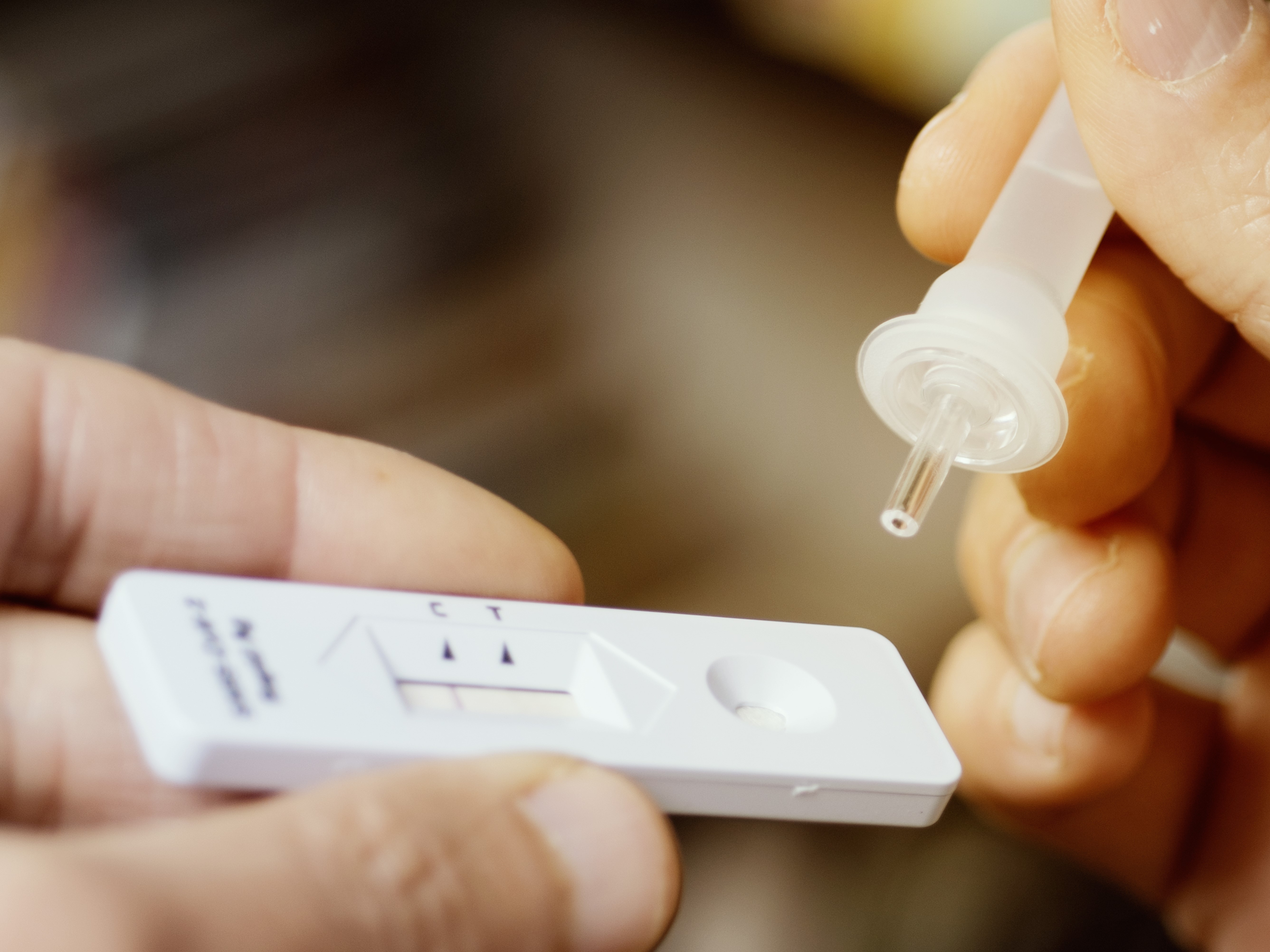 Eine Person hält einen Covid-19-Testkit in einer Hand, während sie mit der anderen Hand eine Tropfpipette über dem Testgerät positioniert. Der Test zeigt mehrere Felder, die zur Anzeige von Ergebnissen verwendet werden.
